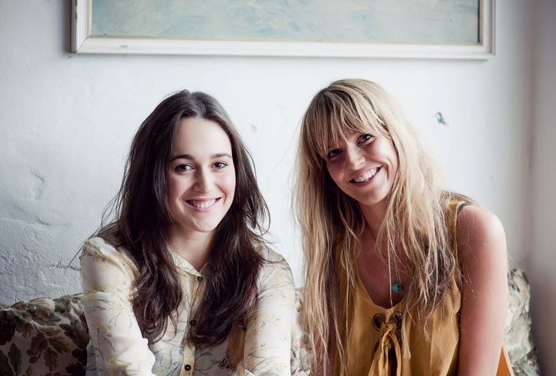 Two friends sitting together and smiling, one with dark hair and one with blonde hair in casual attire.