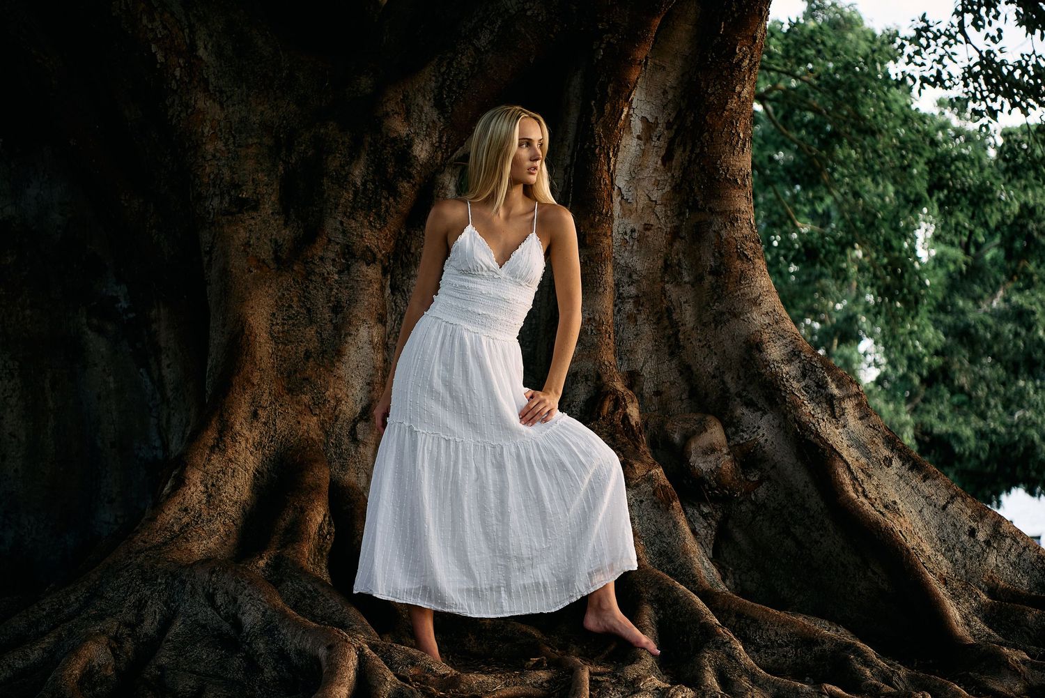 A figure in a flowing white dress poses gracefully among ancient tree roots in a mystical forest setting.