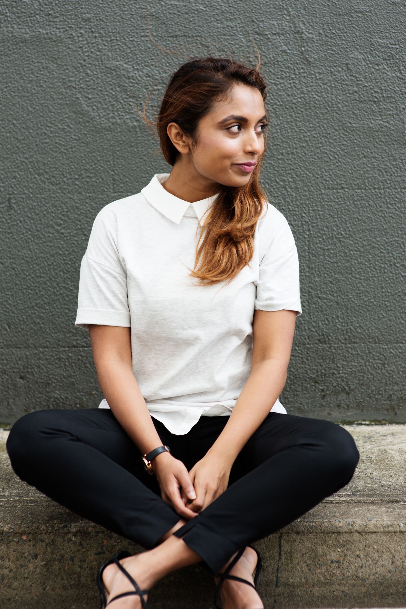 Person in white t-shirt and black pants sitting casually against grey wall.