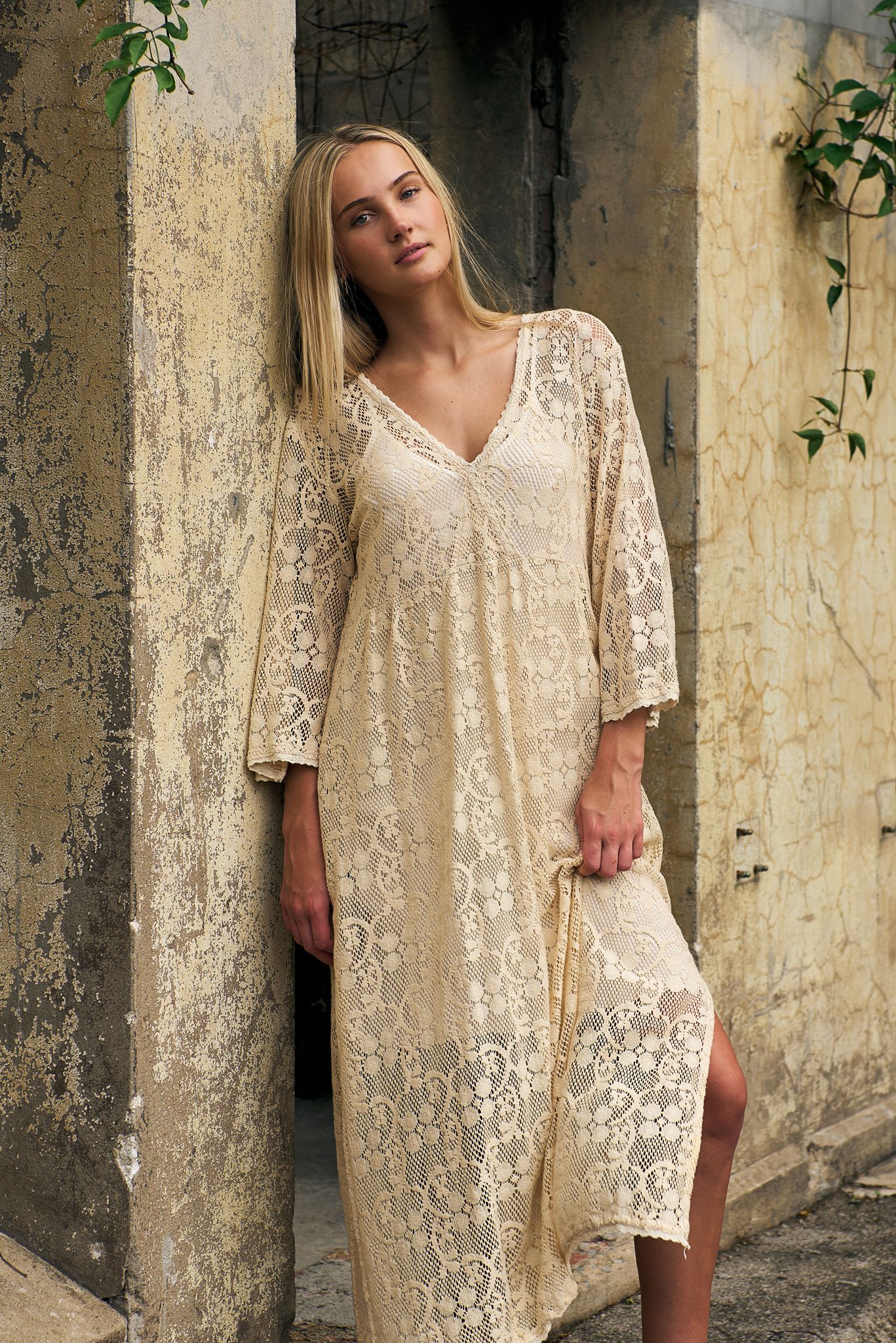 A model in a cream-colored lace shift dress poses against a weathered stone wall with climbing vines.