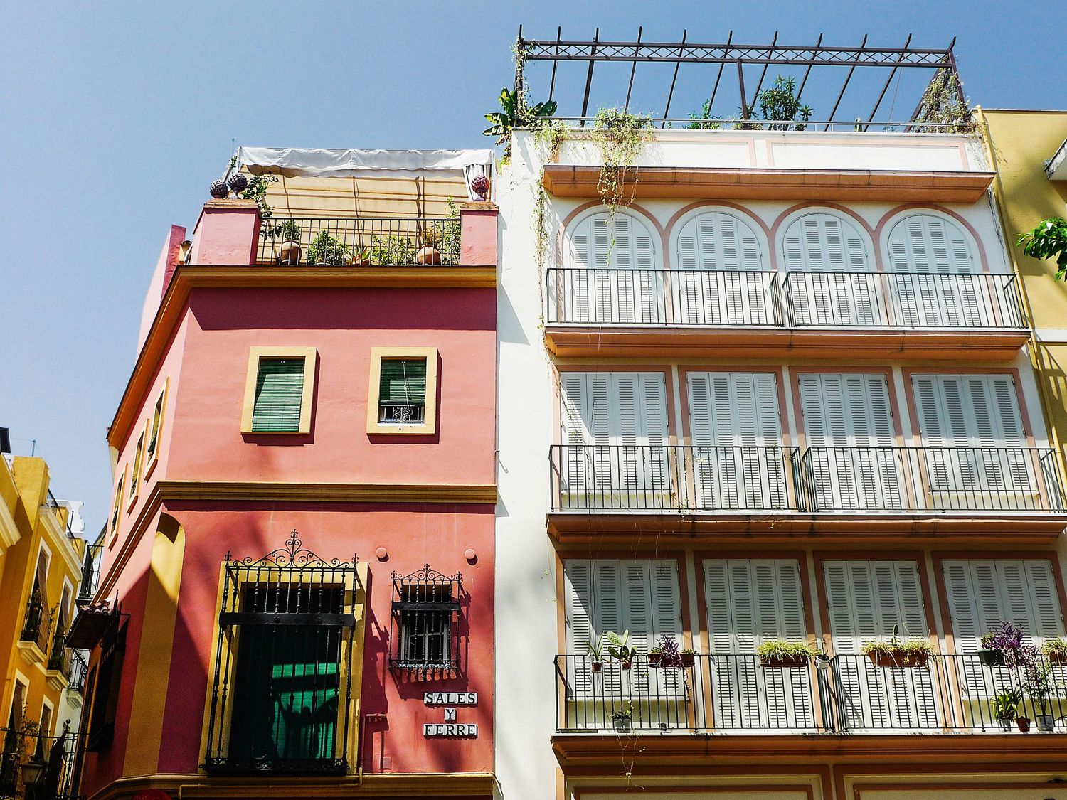 Colorful Mediterranean-style buildings with white balconies and green plants adorning the facades in warm sunlight.