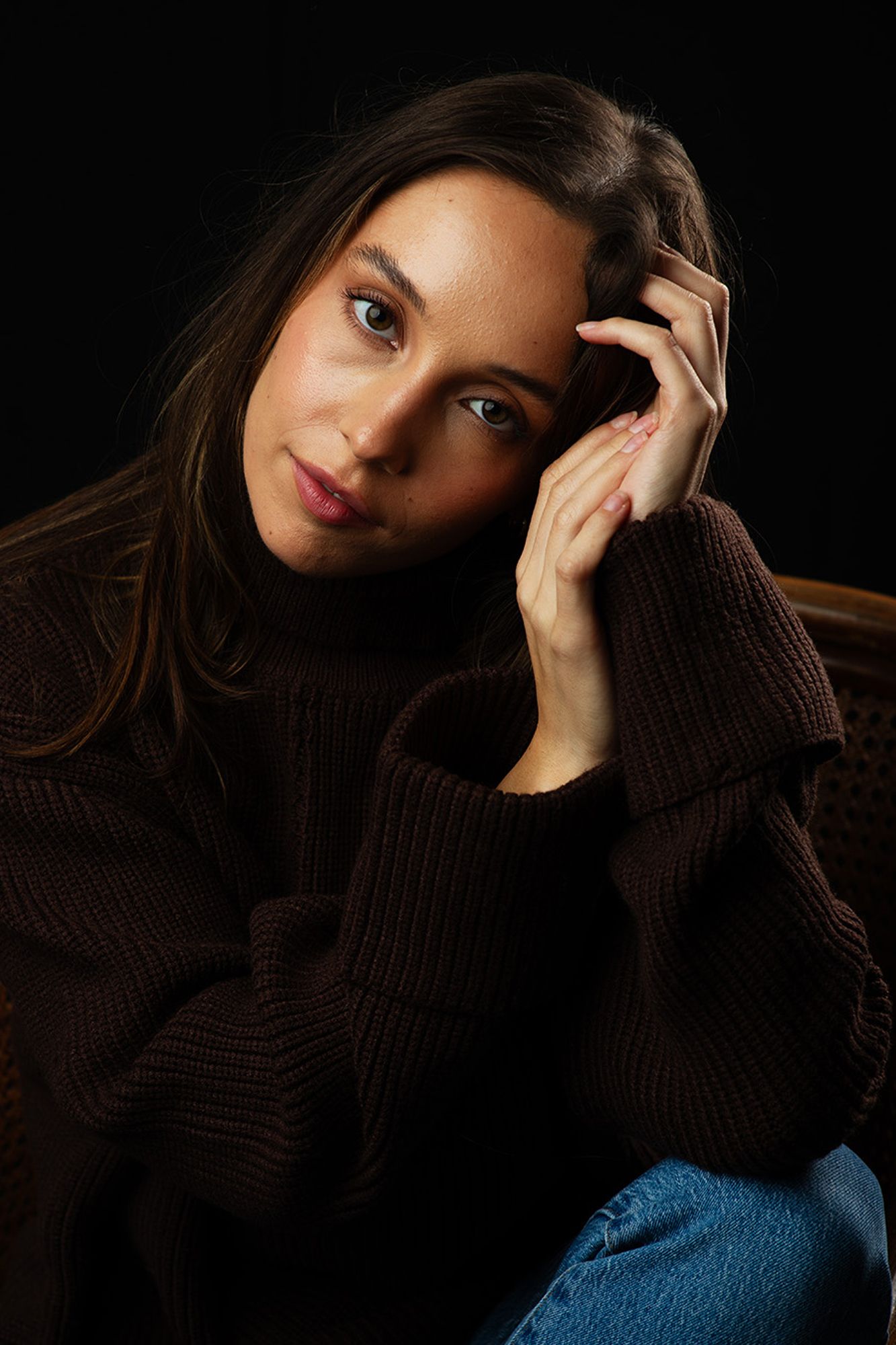 Dramatic studio portrait with warm lighting showing person in brown sweater against dark background.