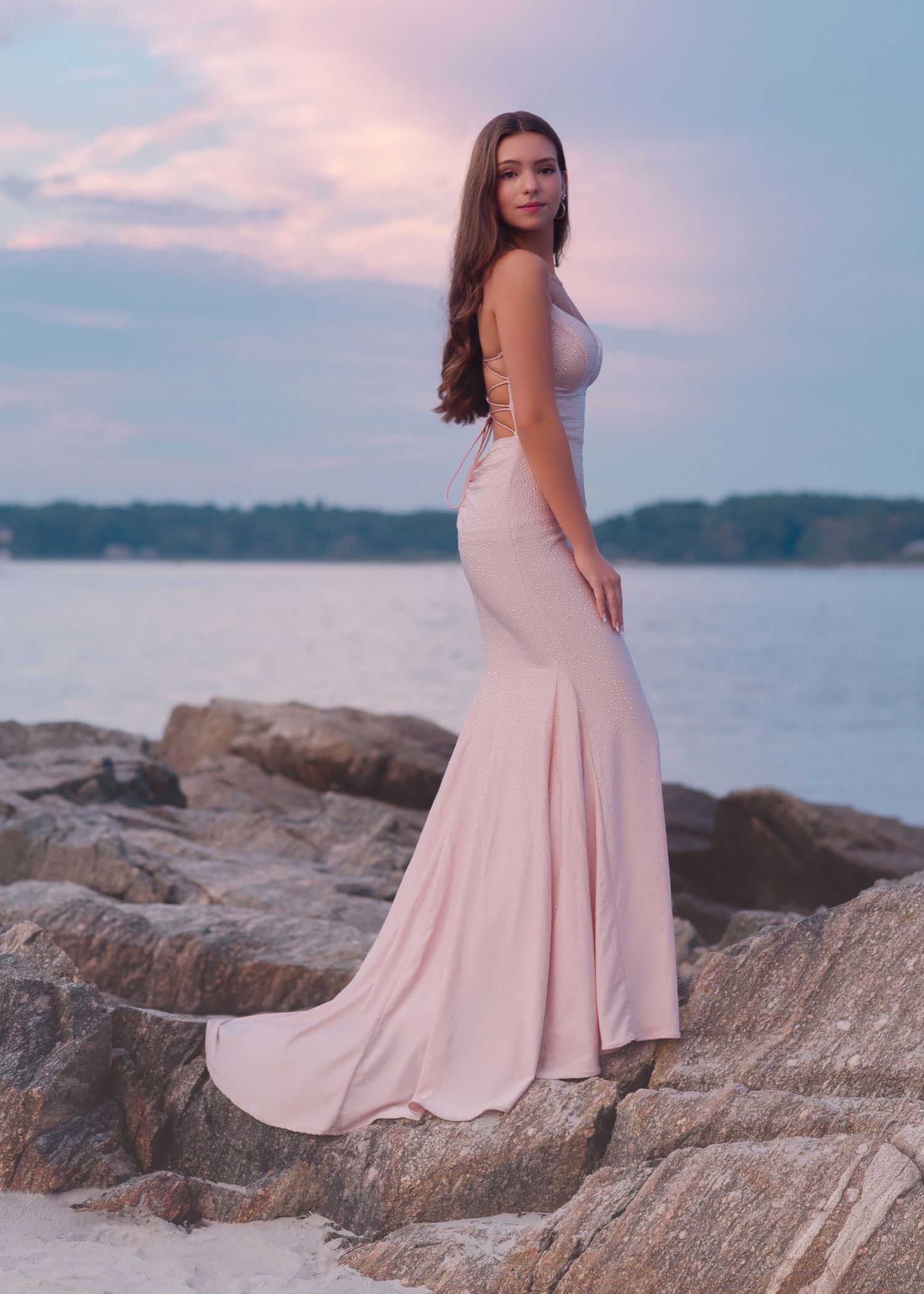A person in a flowing pink dress poses on coastal rocks during sunset at the beach.