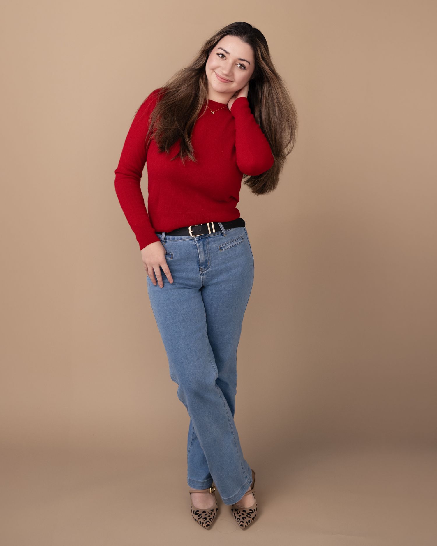 Model in red sweater and blue jeans poses casually against beige backdrop in studio photoshoot.