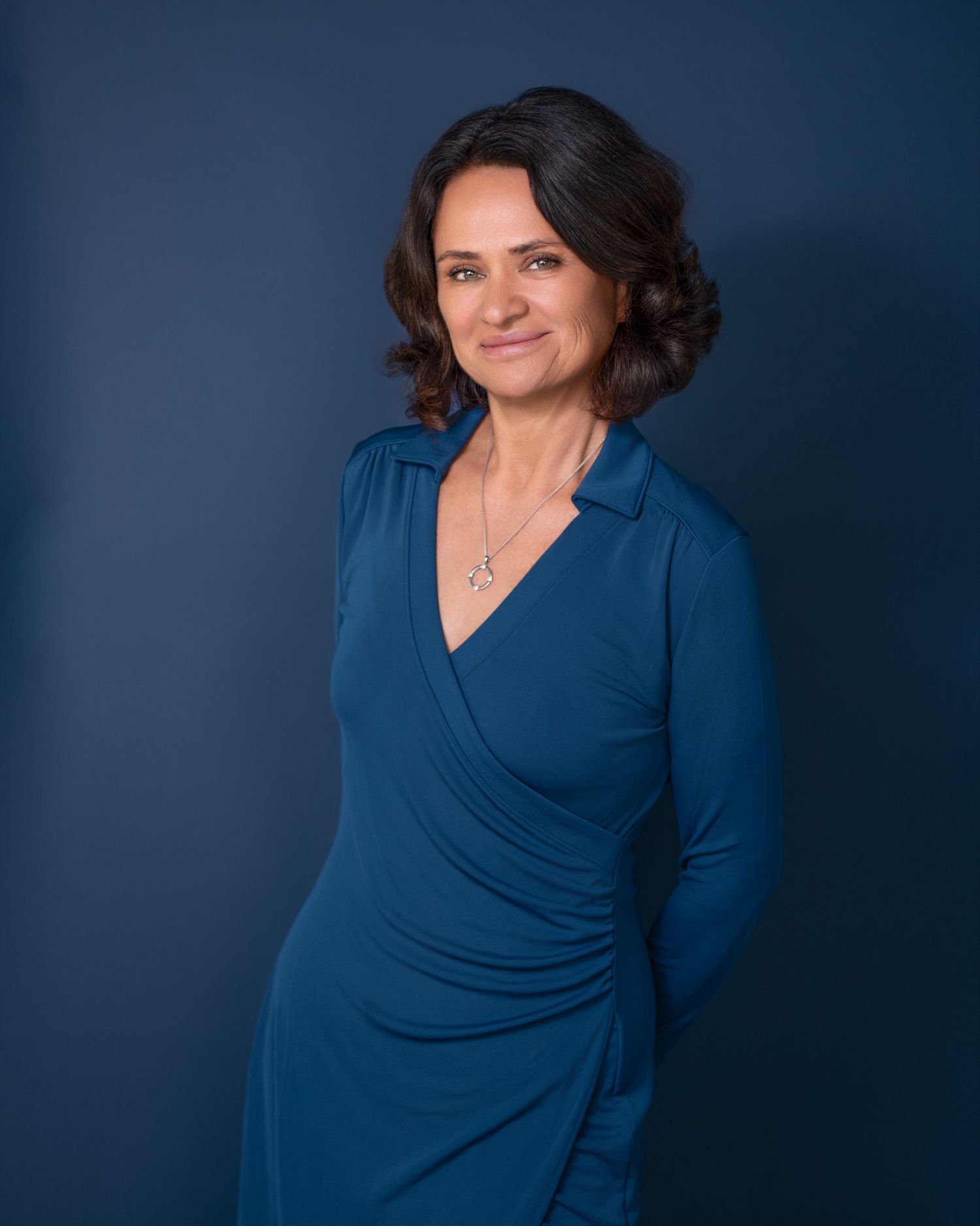 A woman in a teal wrap dress poses confidently against a dark blue background in a professional portrait photo.