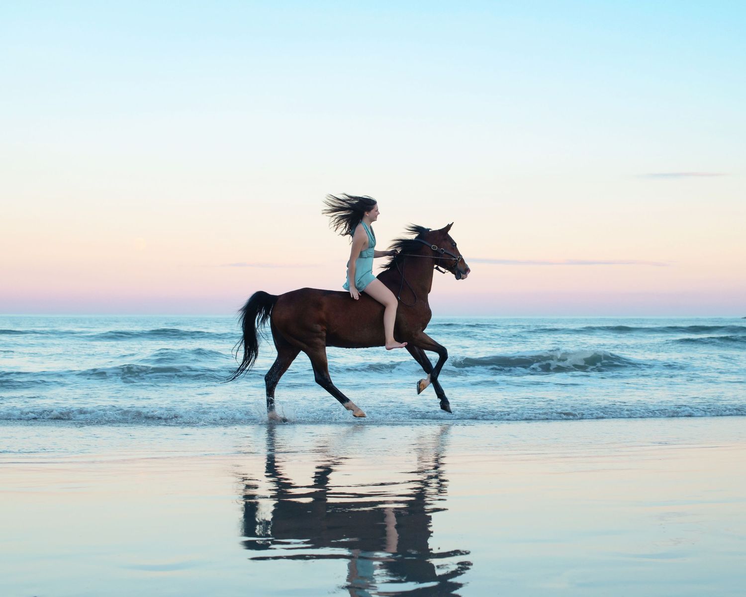 Silhouette of horse and rider galloping on beach at sunset with ocean reflection.