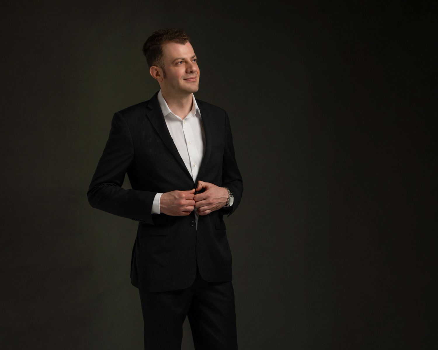 Dramatic portrait of person in black suit adjusting jacket against dark background.