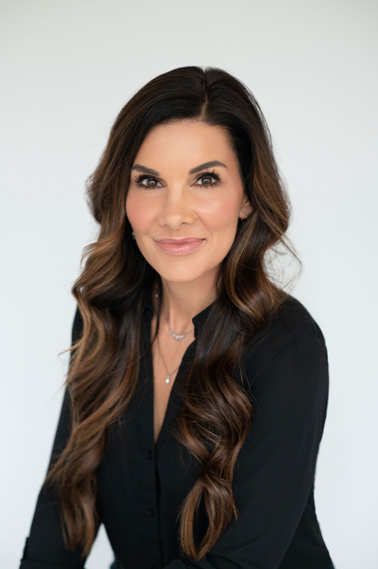 Professional headshot portrait with long wavy brown hair wearing a black blouse against light background.