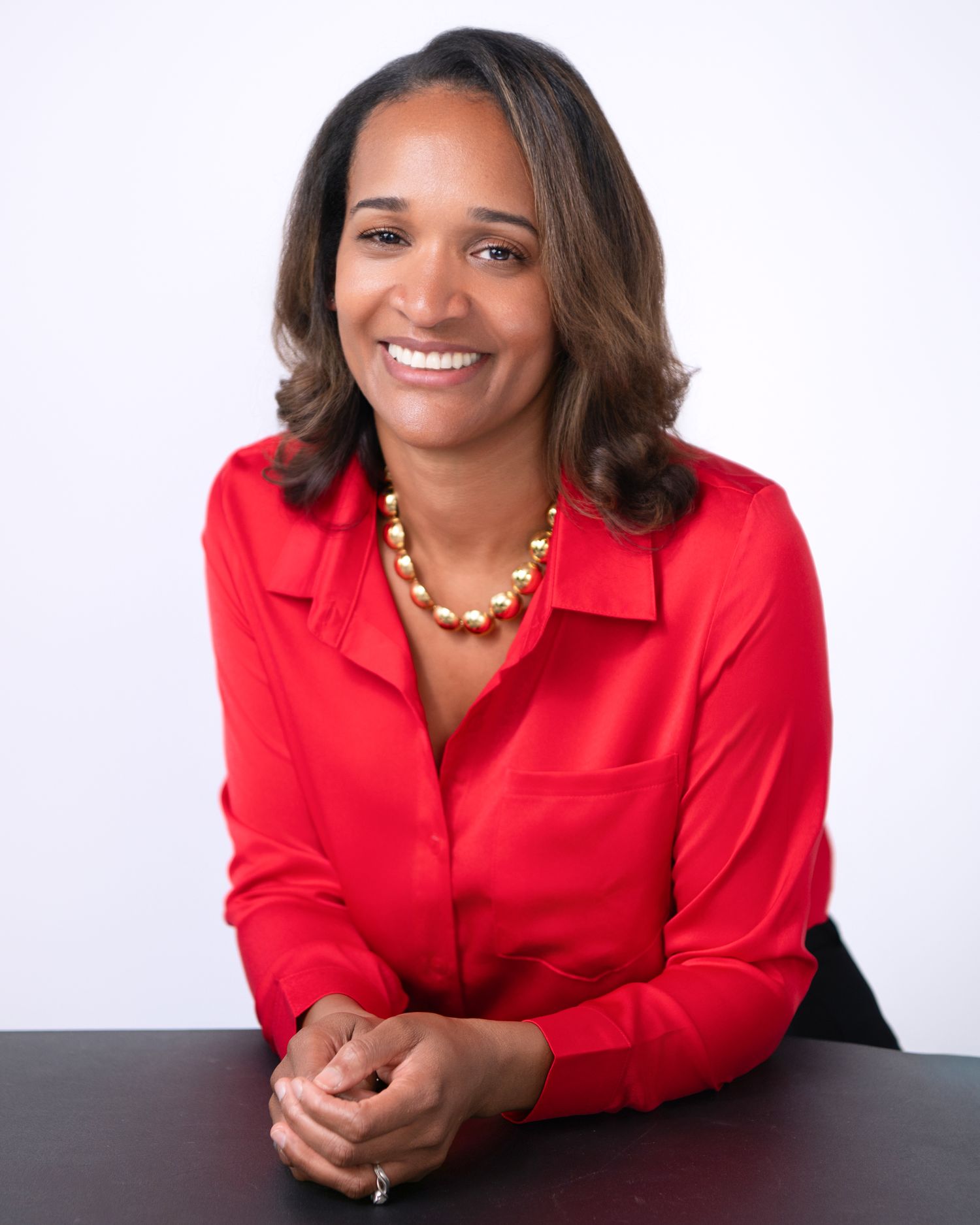Professional woman in red blazer smiling while leaning on dark surface in business headshot series.