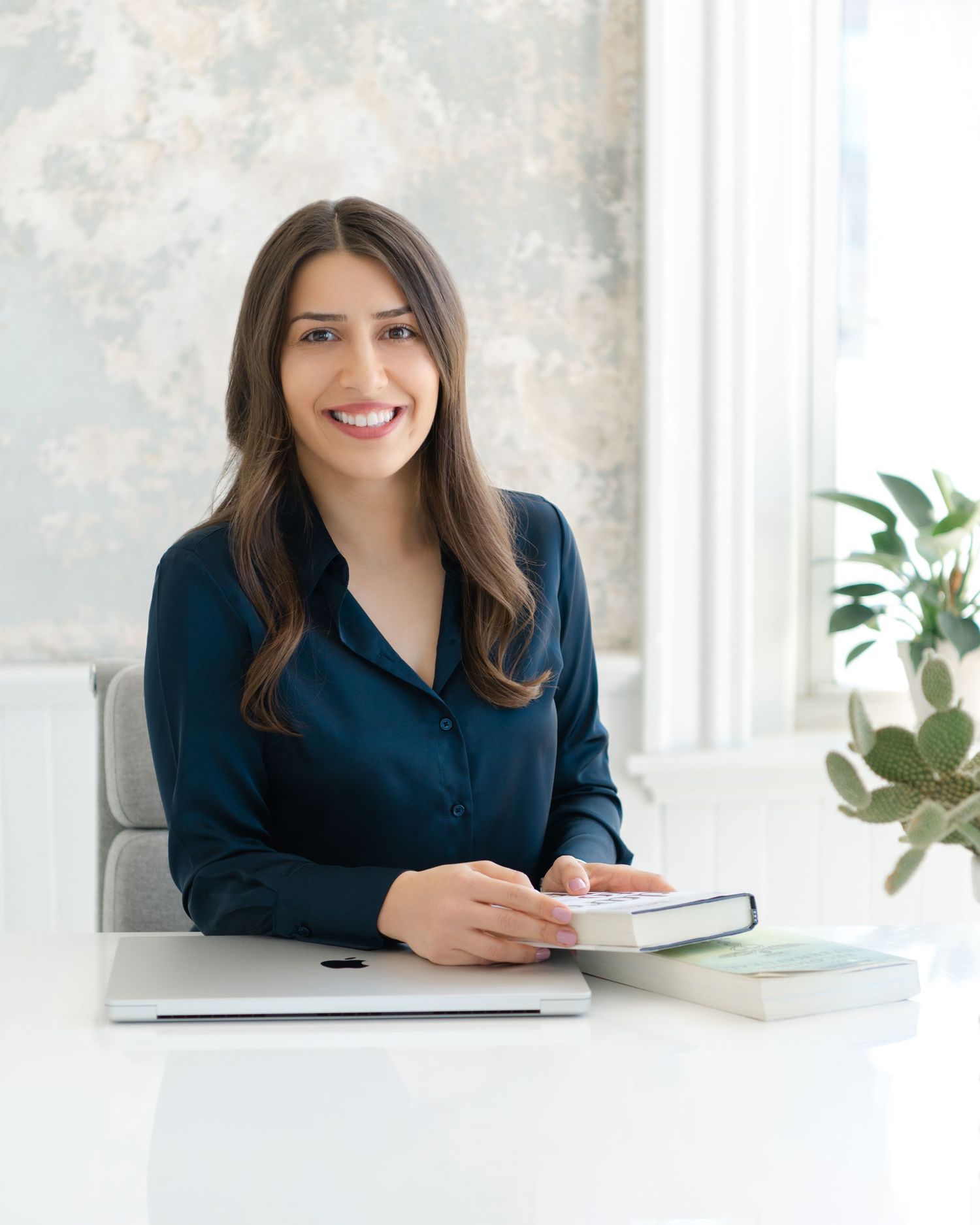 Professional photos of a person in navy blouse working at white desk with plants in background.