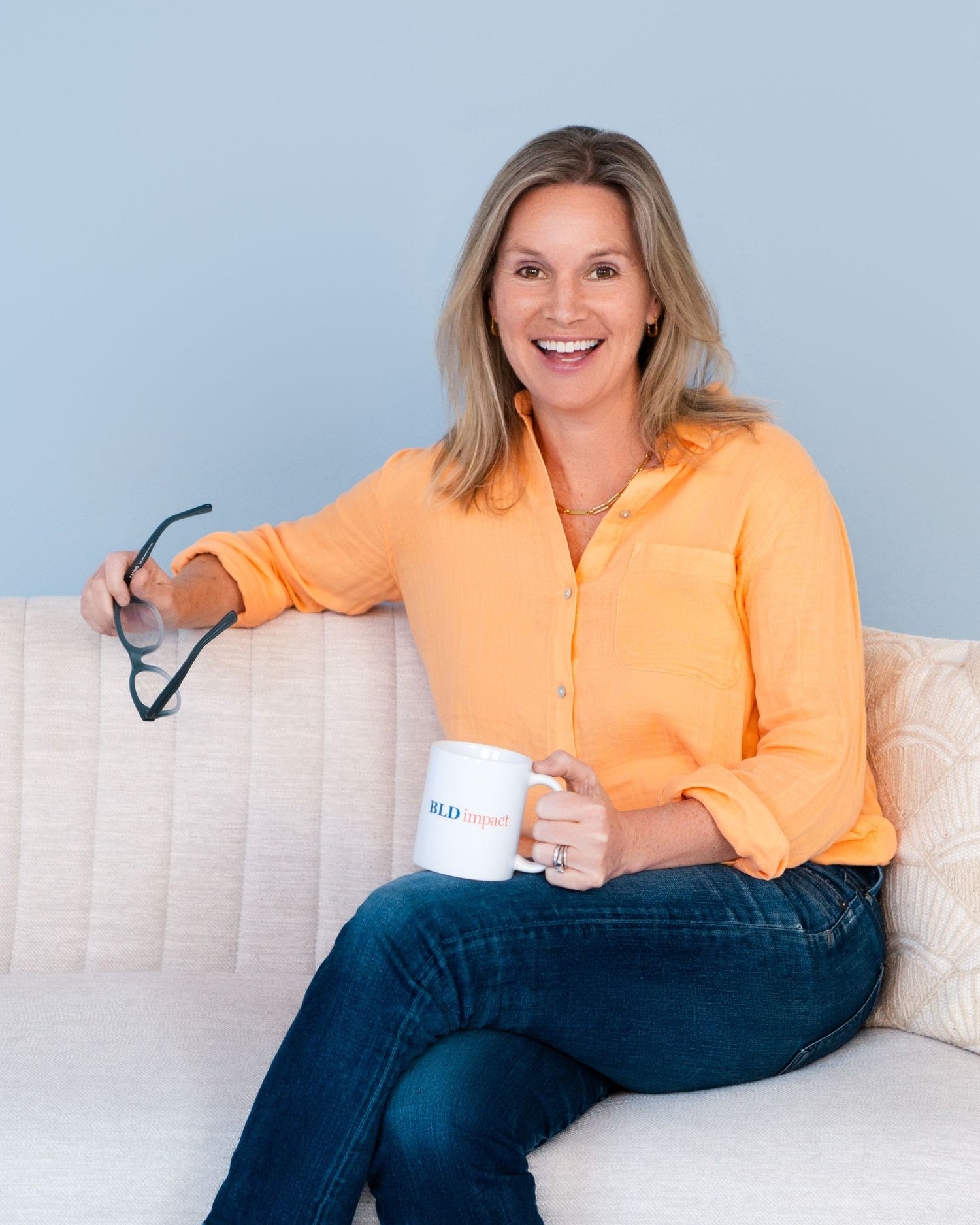 Casual portrait of someone in orange blouse holding coffee mug on cream colored couch.