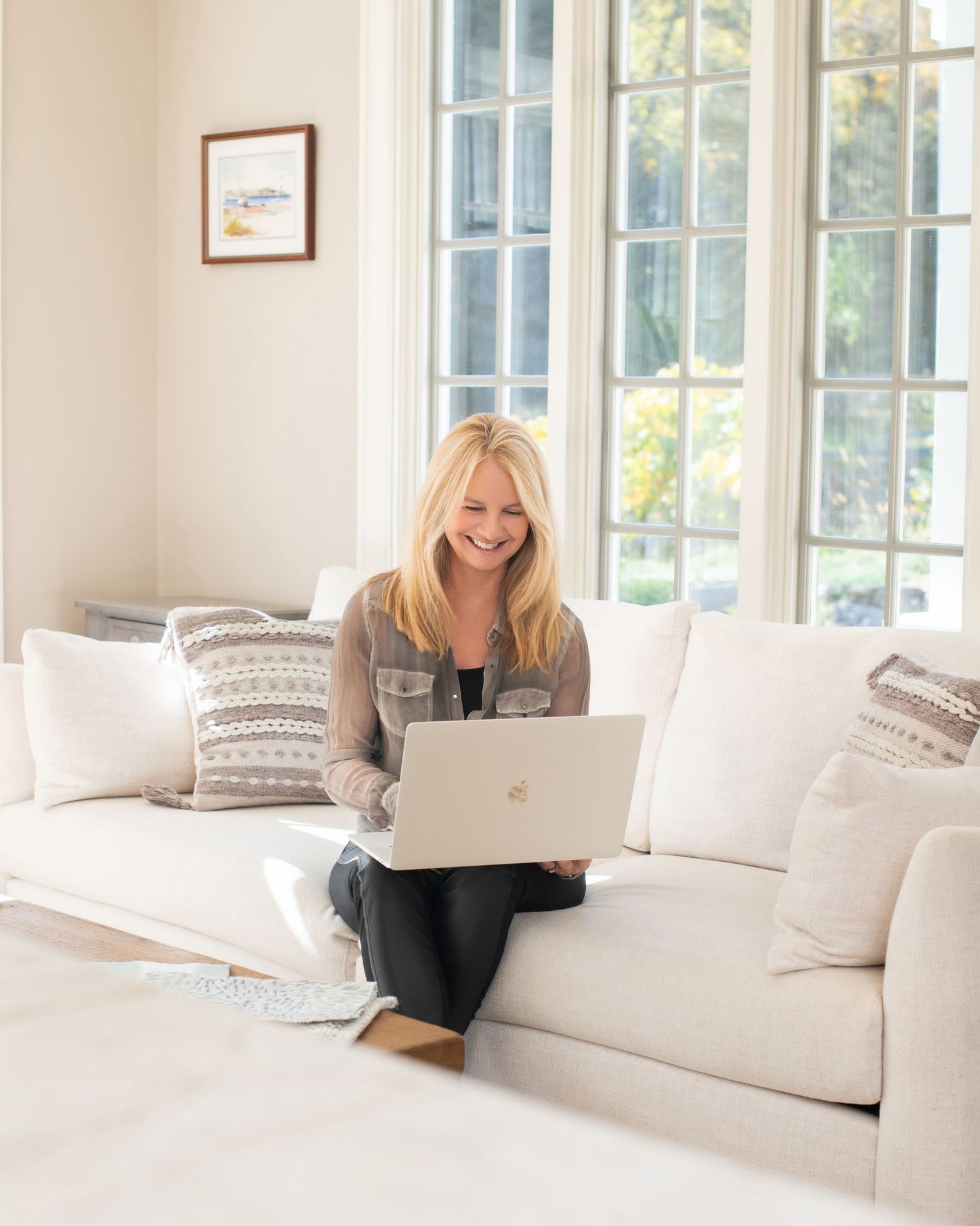 Person working remotely on laptop while sitting comfortably on white couch near bright windows.