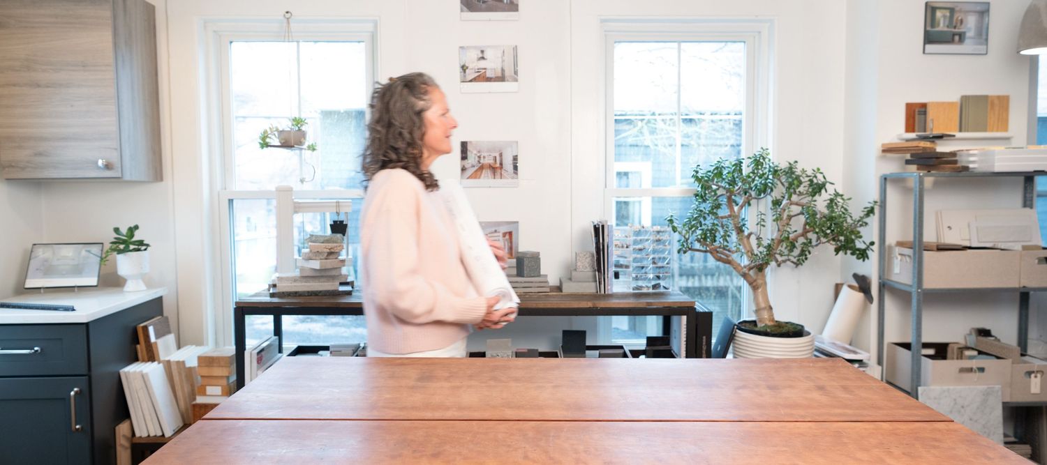 Woman standing at a wooden kitchen island, gazing out bright windows in a modern white kitchen with bonsai tree, in Kittery, ME