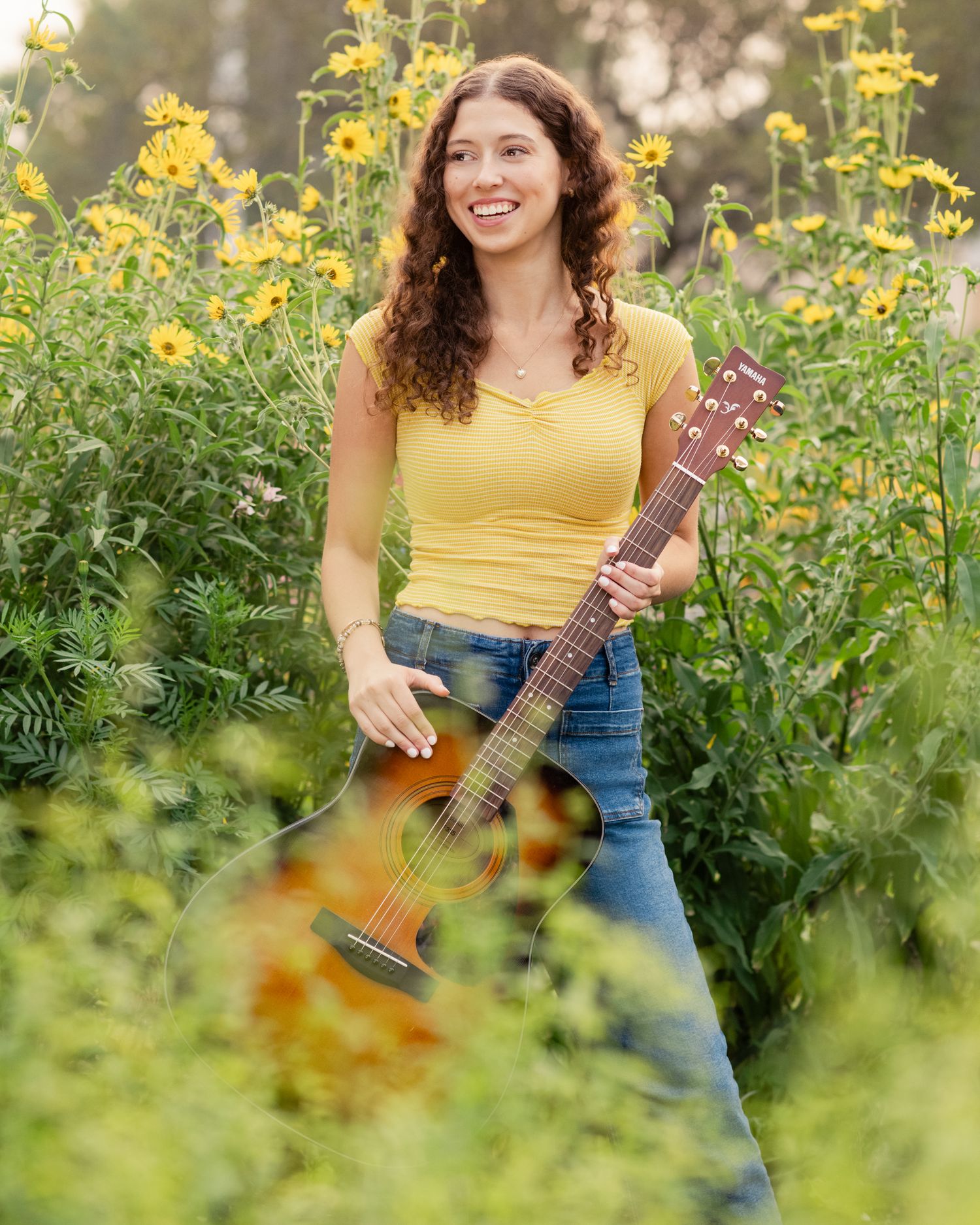 Woman in yellow top playing ukulele among blooming yellow wildflowers in a garden.