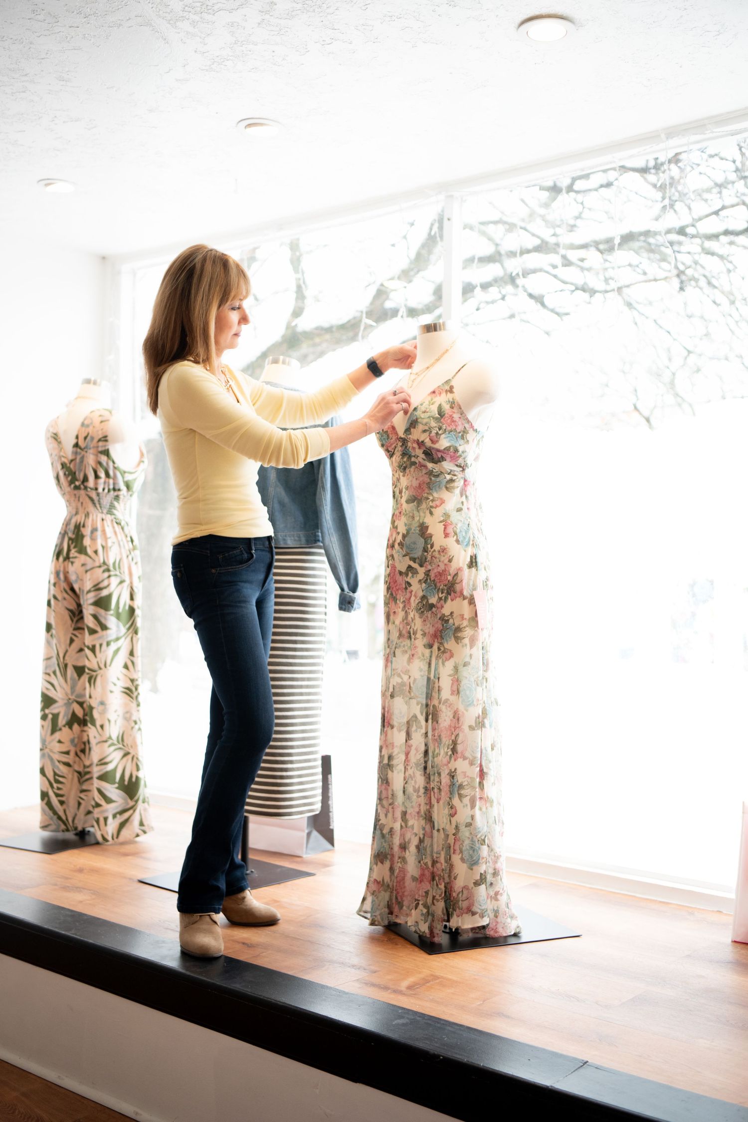 A designer adjusts a floral gown on a dress form mannequin in a bright boutique with large windows for a Branding shoot in Amesbury, MA