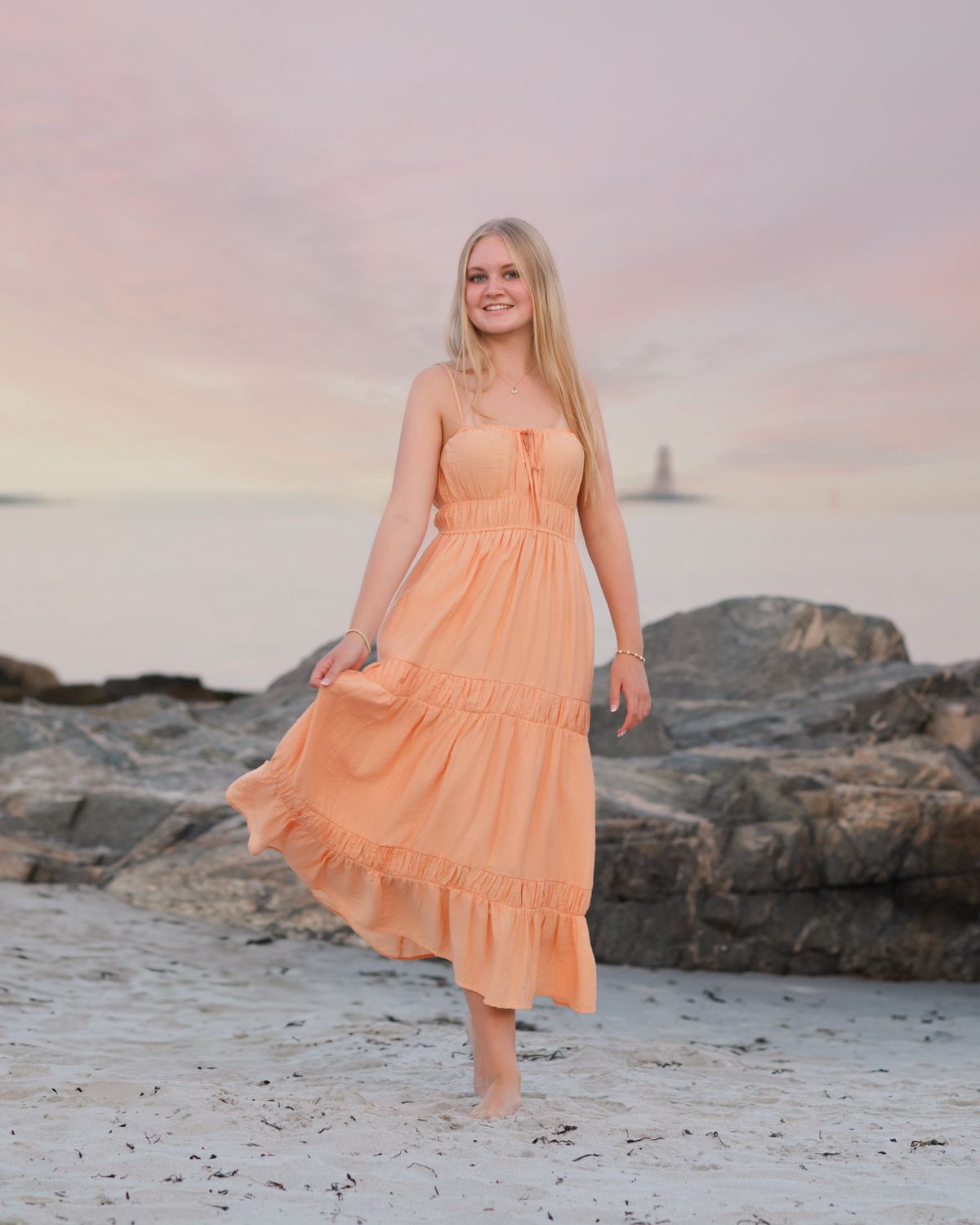 Beach photoshoot featuring flowing peach dress against coastal rocks at sunset.