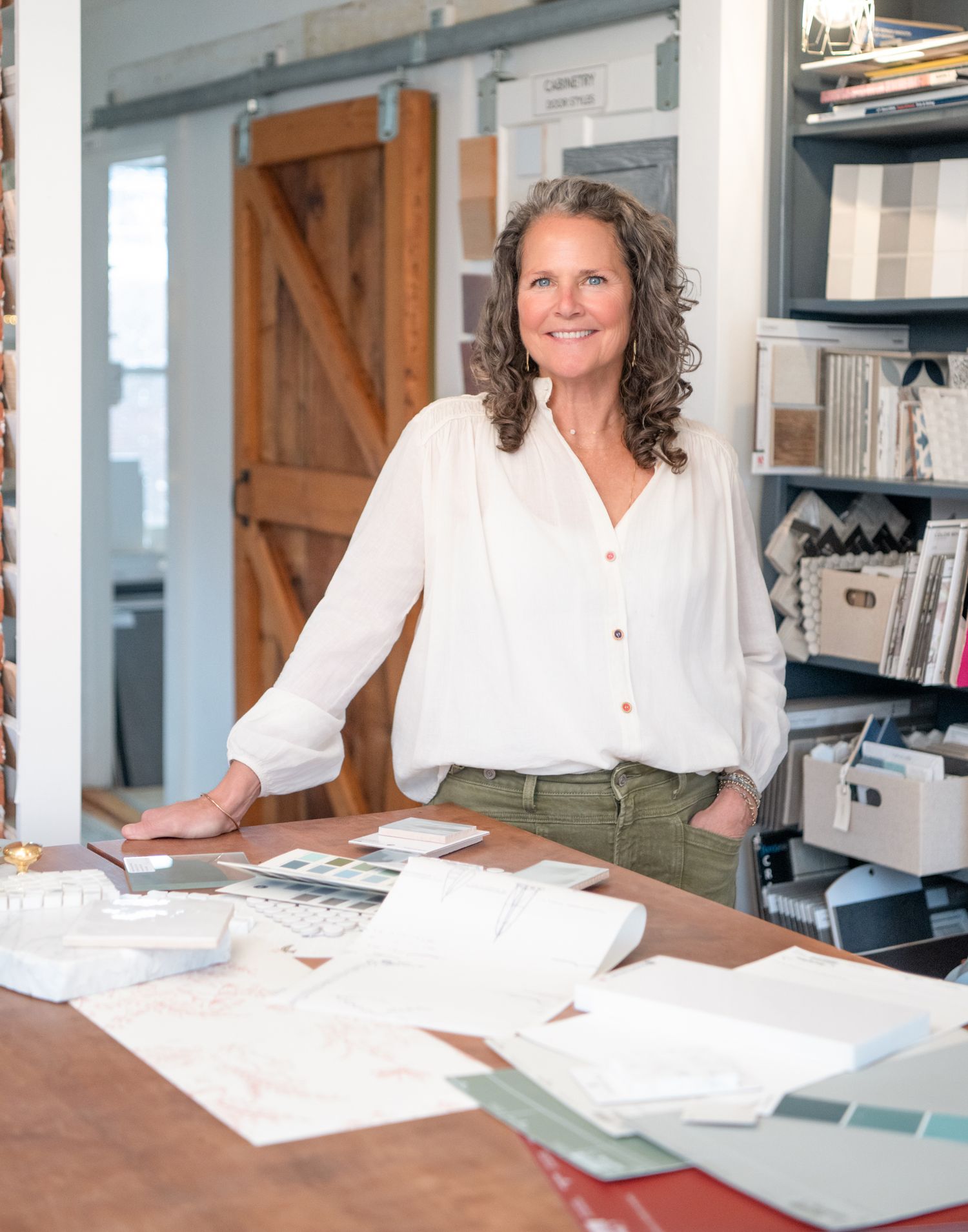 Woman interior designer in Kittery ME, smiling at desk with fabric samples and design materials in a modern studio workspace.