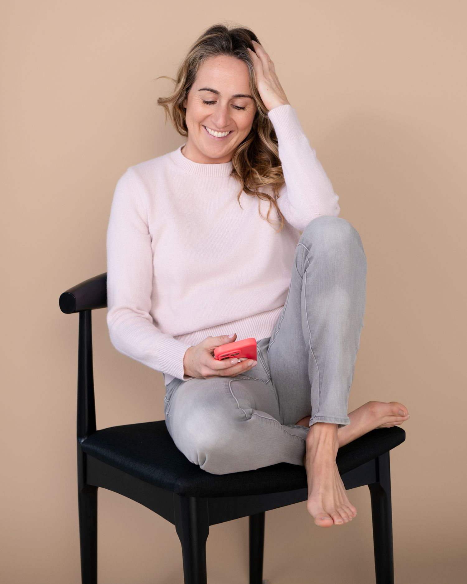 Person in pink sweater and grey pants sitting casually on black chair while checking phone.