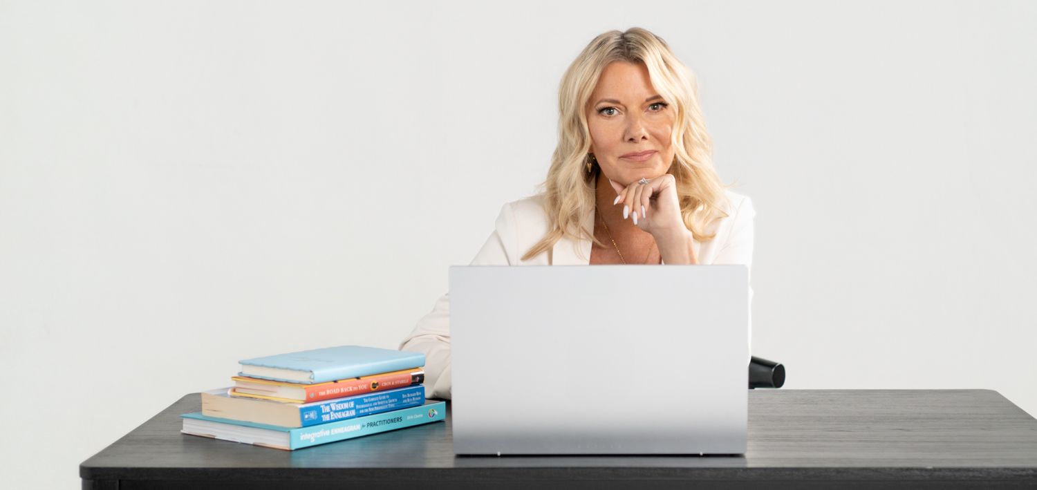 A person wearing a pink sweater sits at a desk with books and a laptop while working from home in Amesbury, MA.