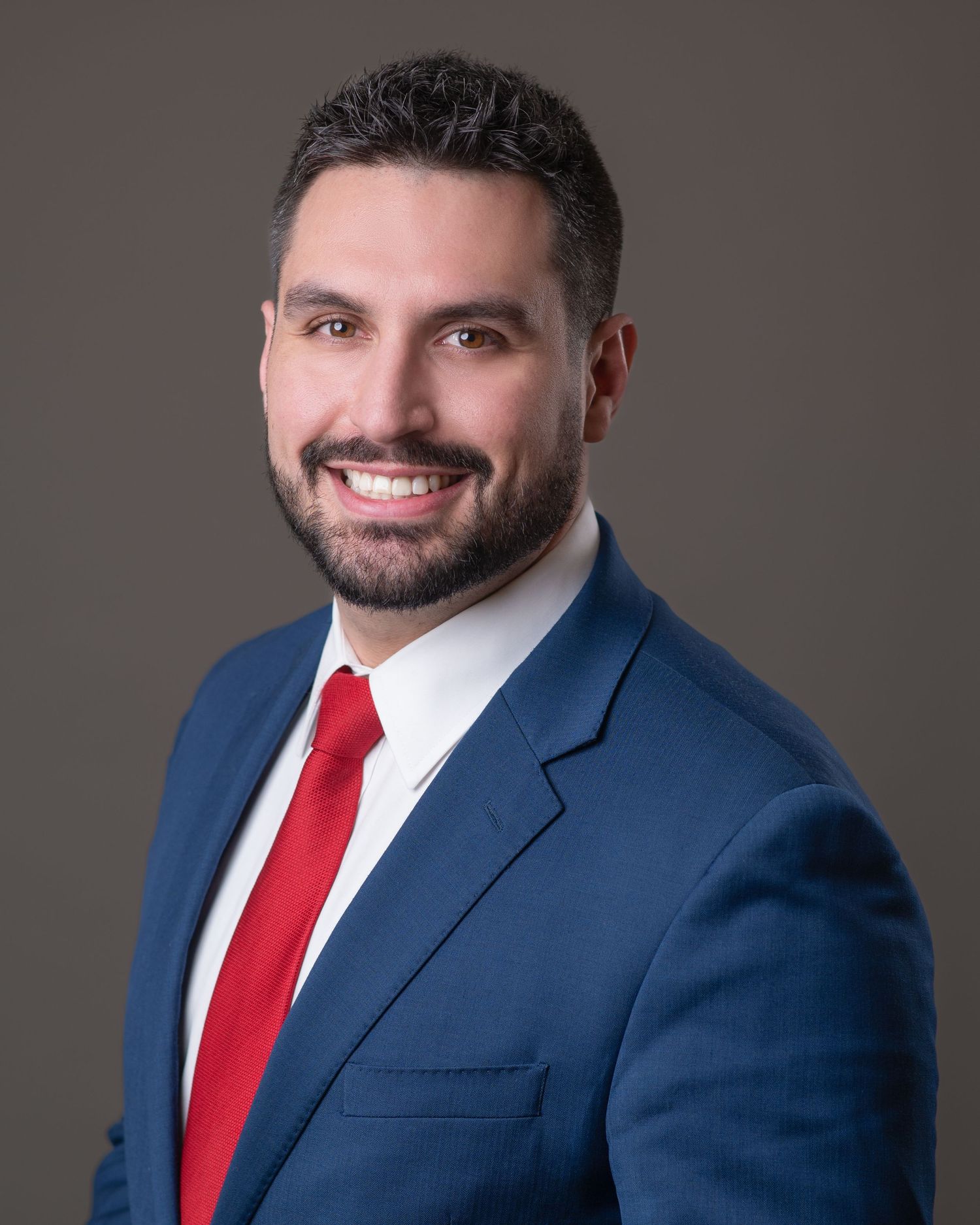 Professional headshot of someone wearing a navy blue suit and red tie against a gray studio backdrop.