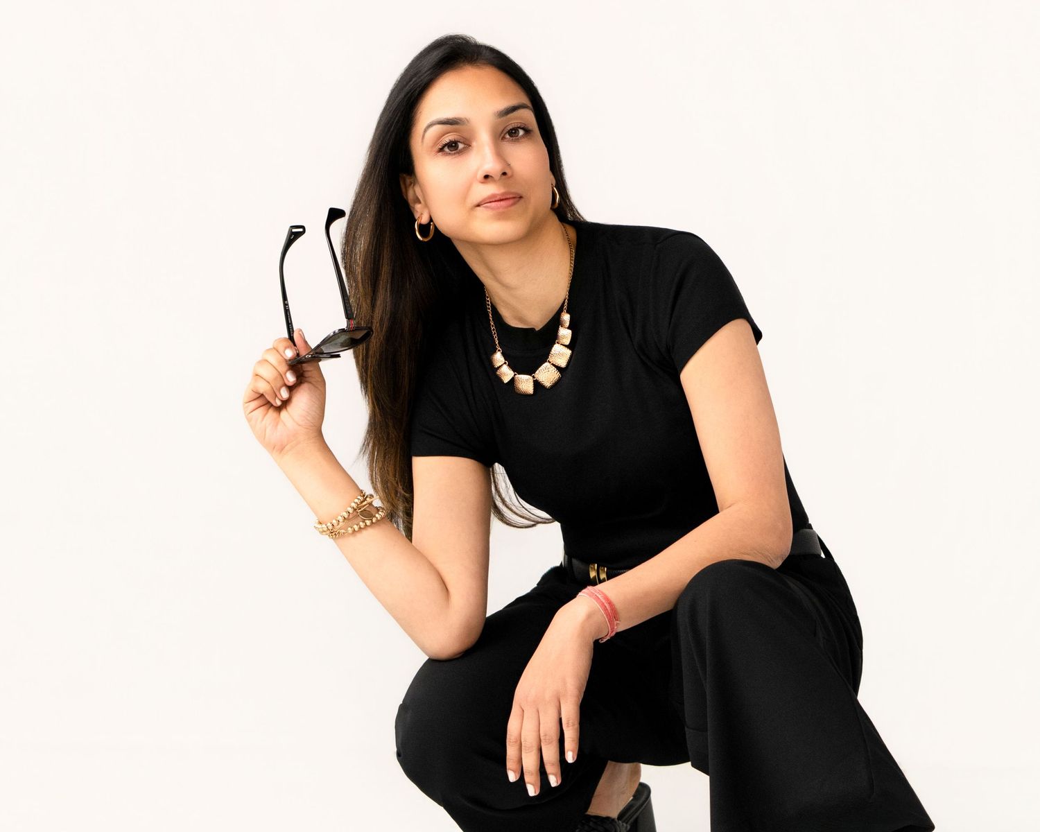 Woman in black outfit holding glasses, wearing gold jewelry, posed confidently against white background, Amesbury, MA