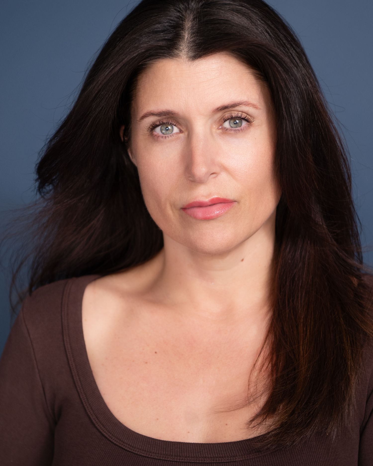 Professional headshot of a woman with long dark hair wearing a brown top against a blue background.