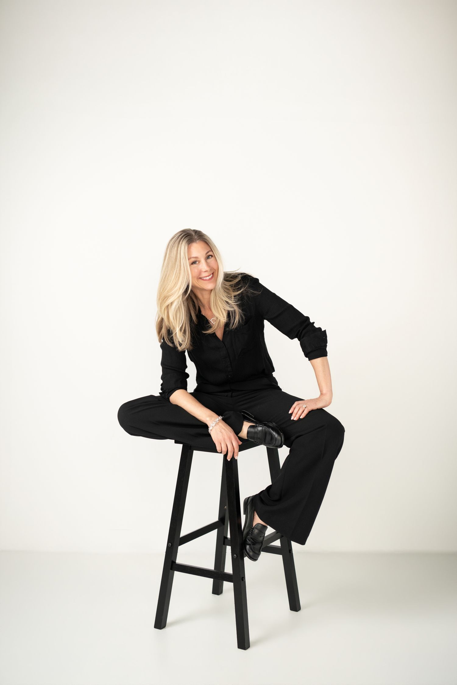 Blonde woman in black outfit sitting on a wooden stool against a white background, smiling confidently.