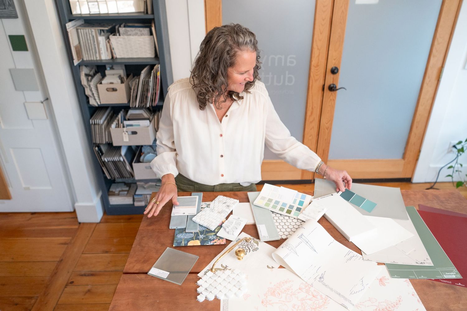 Woman reviews interior design samples and color swatches spread across a wooden table in a bright home office in Kittery, ME.