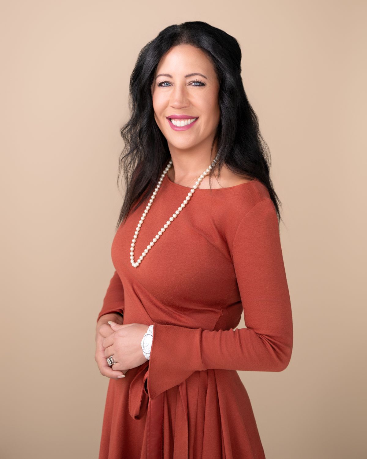 Woman in rust-colored dress with pearl necklace poses against beige backdrop.