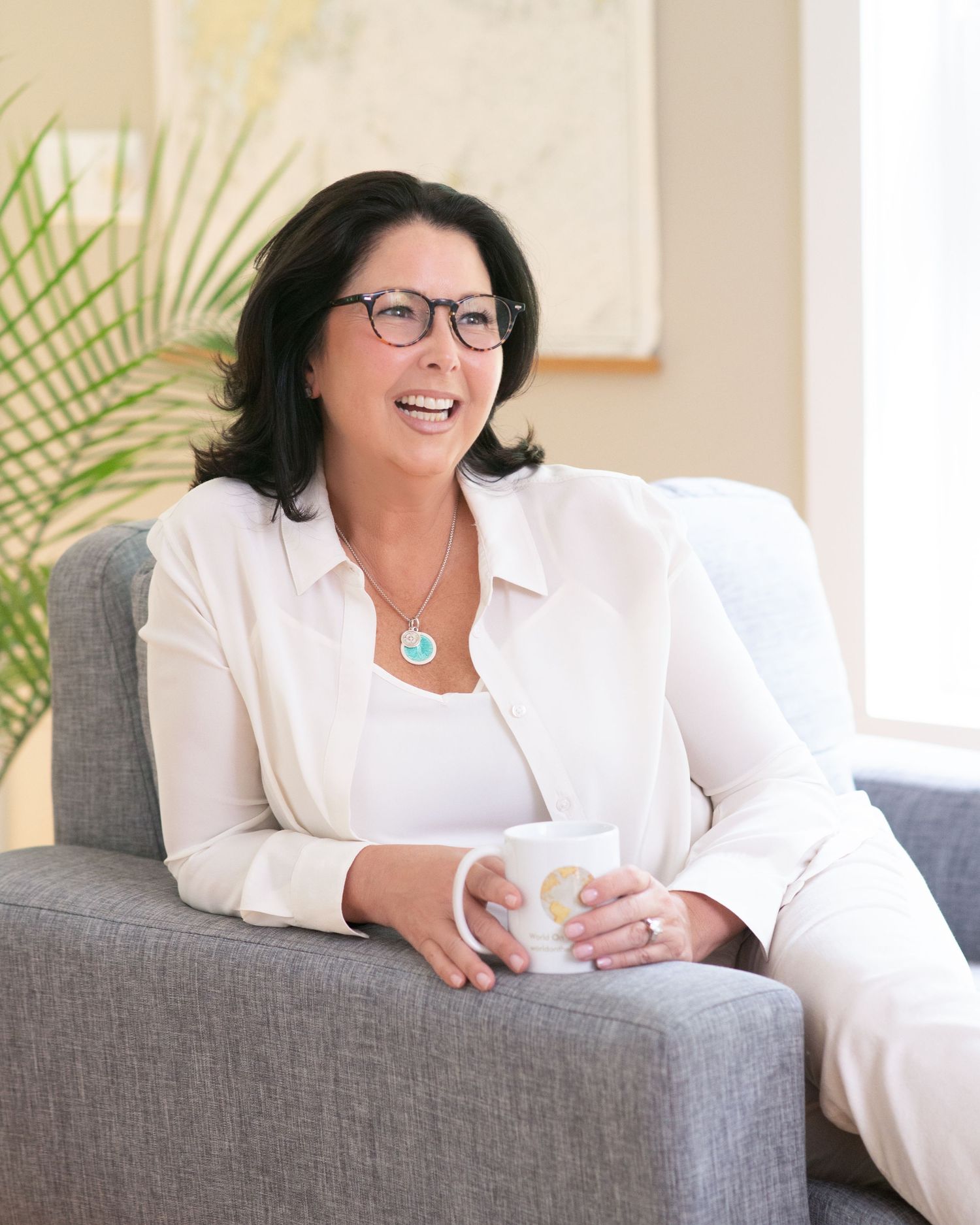 Professional portrait of someone in white blazer sitting on grey chair with coffee.