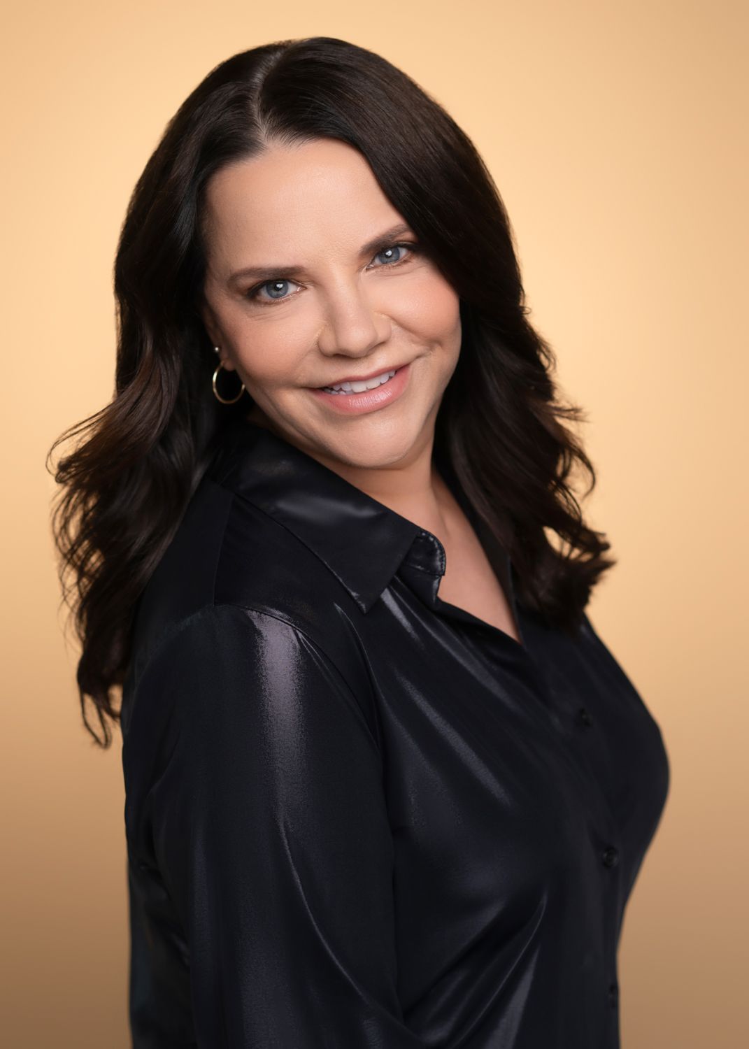 Studio portrait of person in black silk blouse against warm peach background.