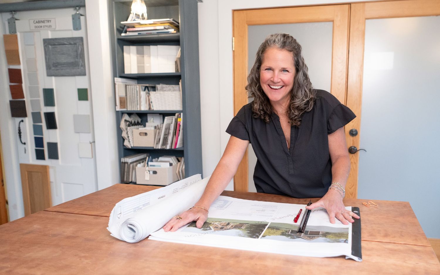 Woman in Kittery, ME, smiling at desk with architectural plans and design samples in a bright interior design studio.