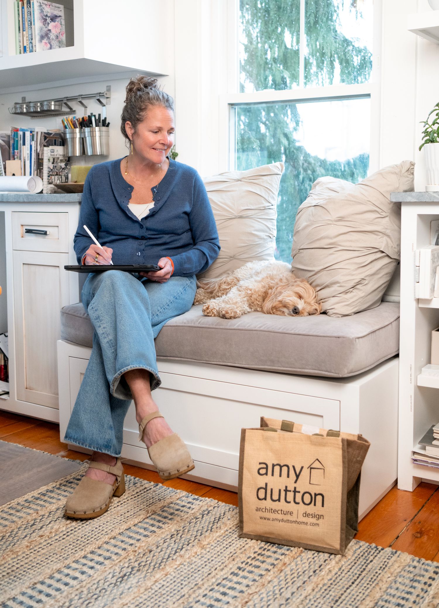 Amy Dutton sits on a cozy built-in bench with her fluffy dog, writing in a bright home studio space in Kittery, ME.