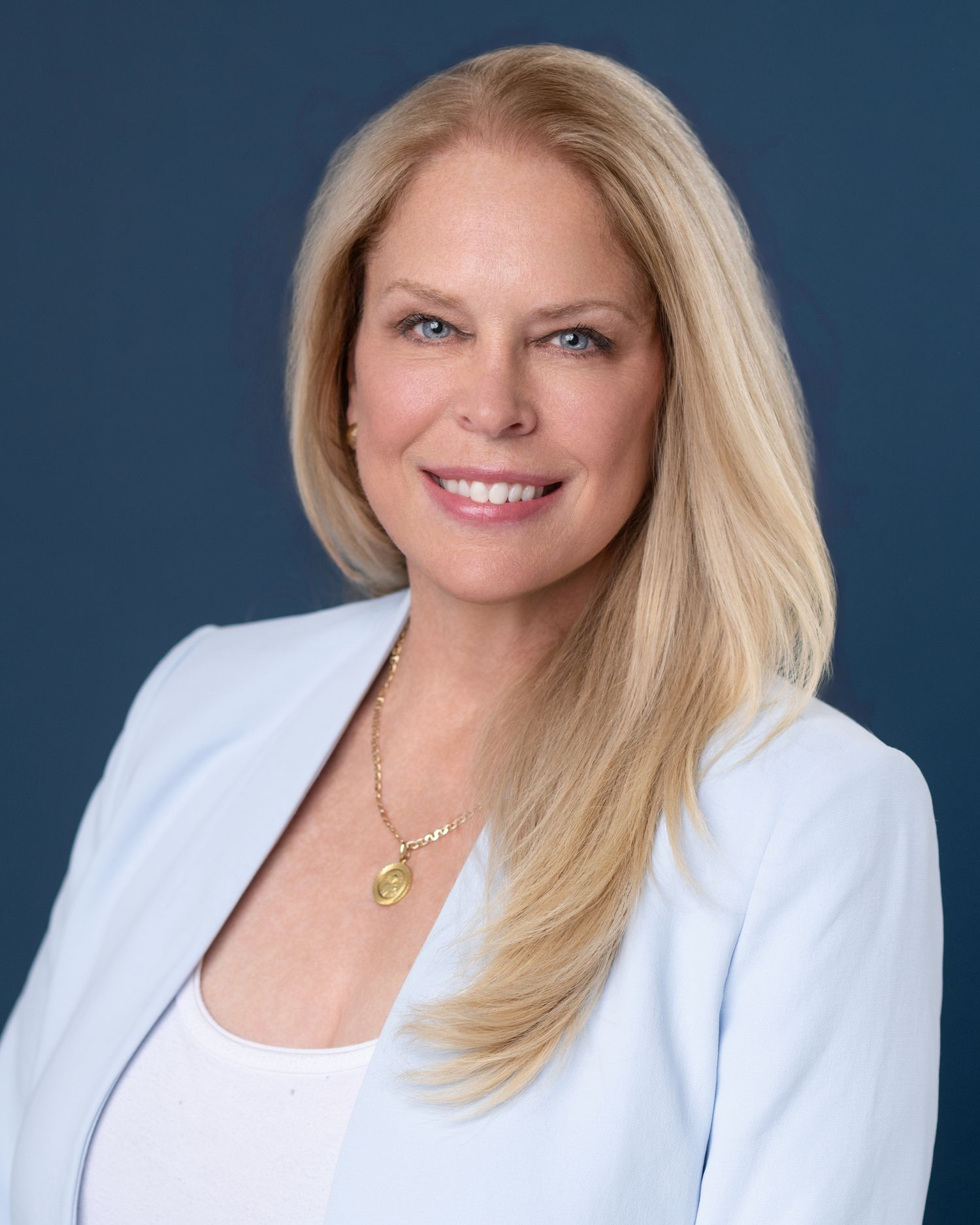 Professional headshot of a woman in white blazer with blonde hair against blue background.