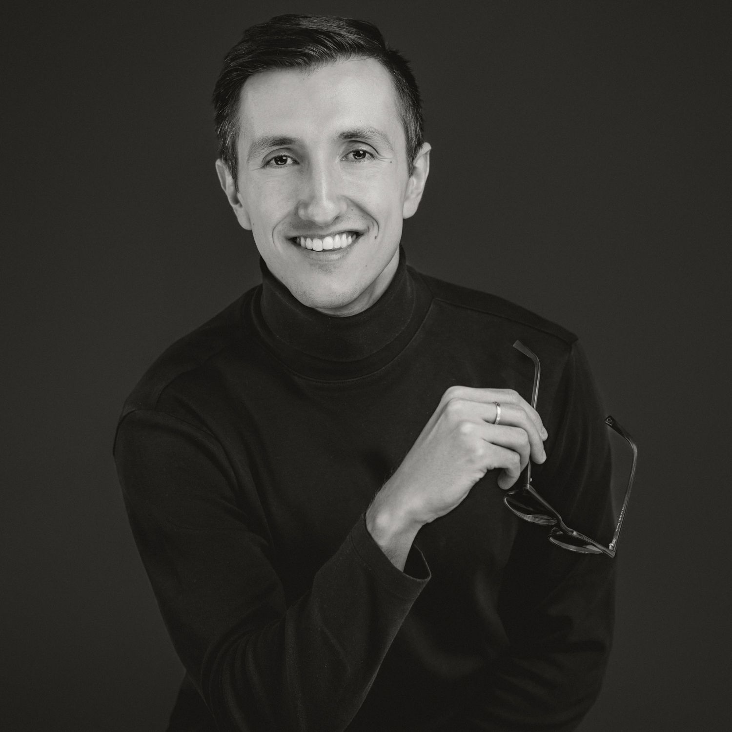 Professional headshot in black turtleneck against dark background.