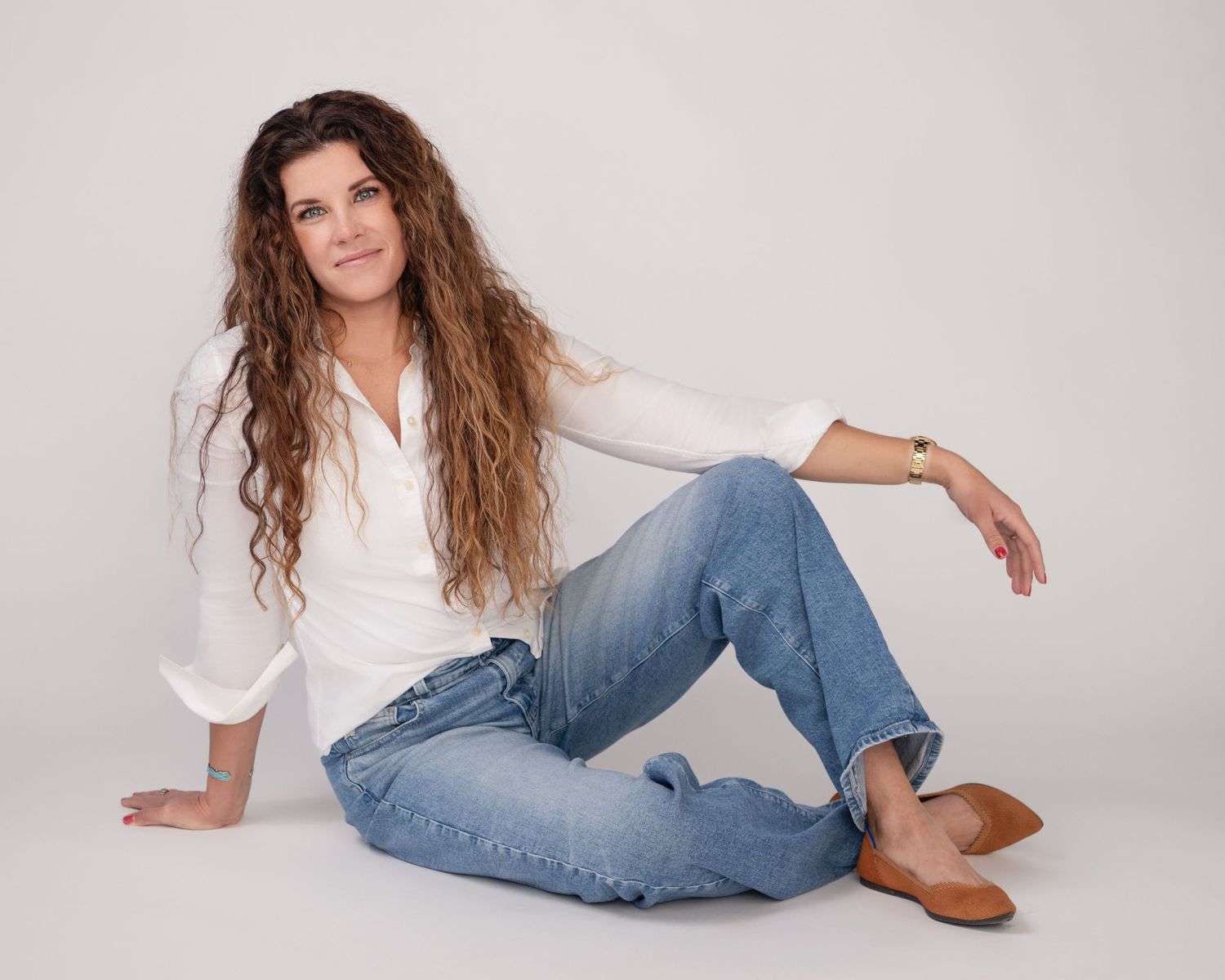Person with long wavy hair wearing white button-up shirt and distressed jeans poses casually on white background. Amesbury, MA