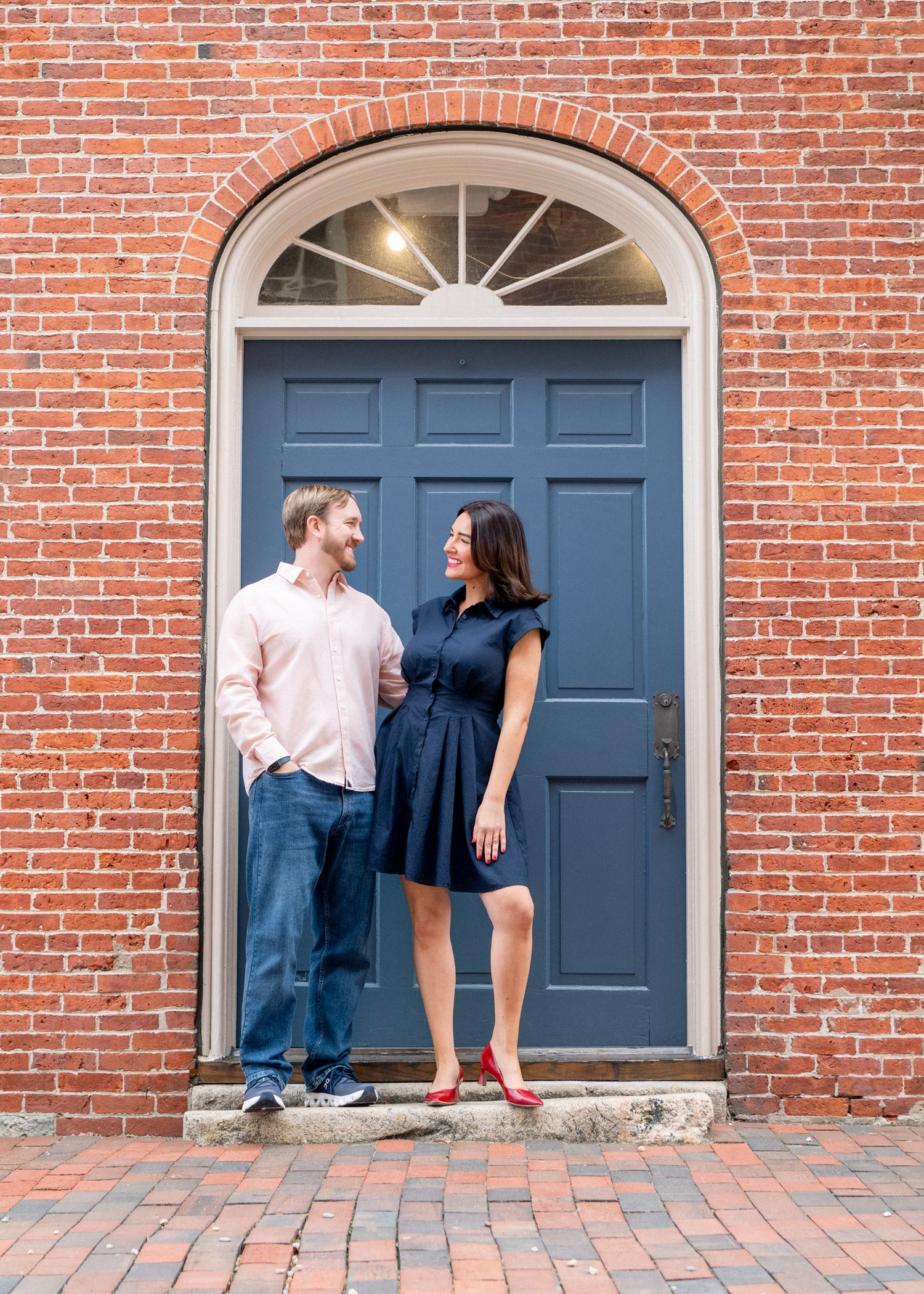Couple smiling at each other in front of a blue arched door on a red brick building, woman in navy dress and red flats.