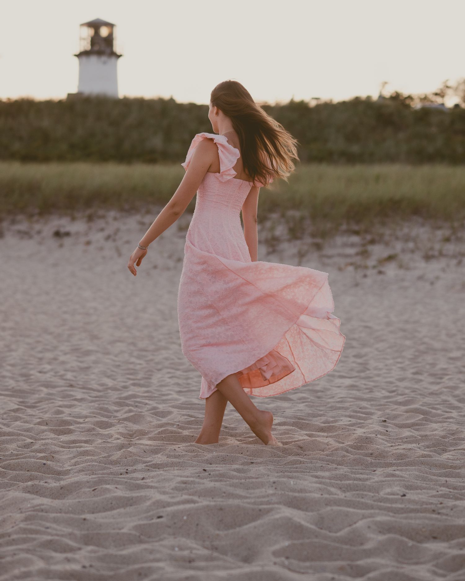 Flowing pink dress twirls on beach with lighthouse silhouette at dusk.