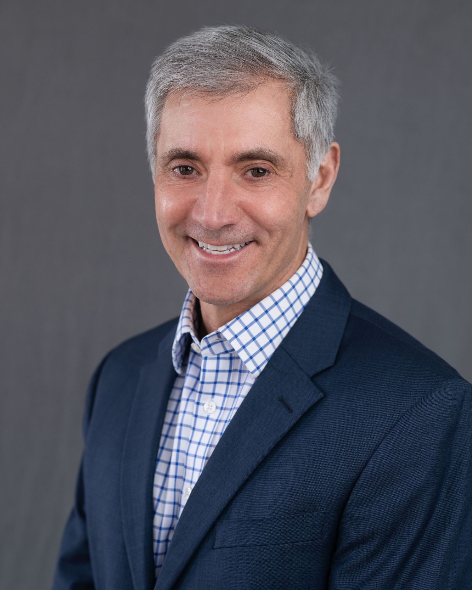 Professional headshot of person in navy blazer and checkered shirt against gray background.