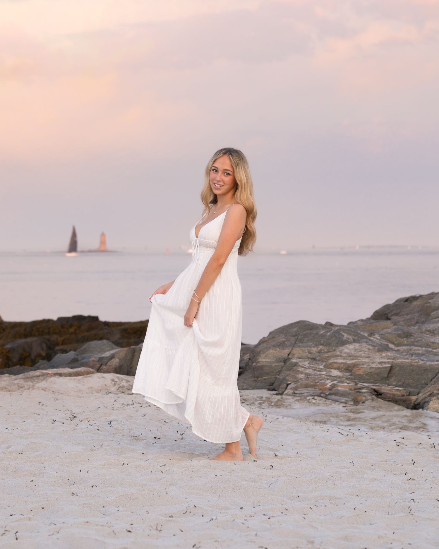 Seaside portrait in flowing white dress on rocky beach at sunset.