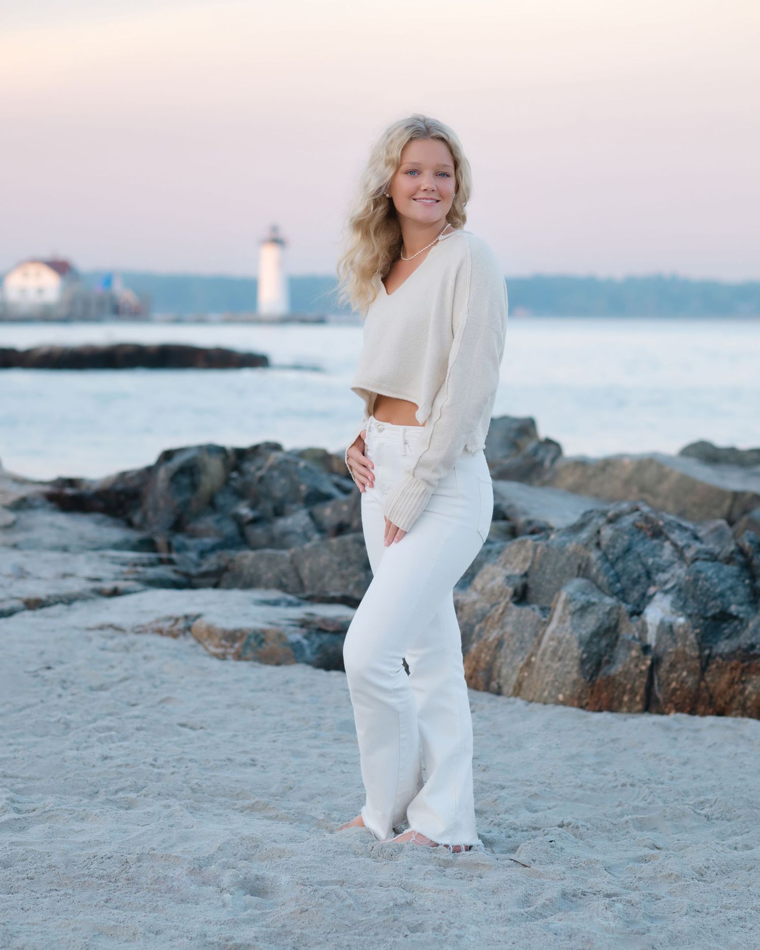 Woman in all white outfit poses gracefully on sandy beach with rocky coastline and lighthouse in background at sunset.