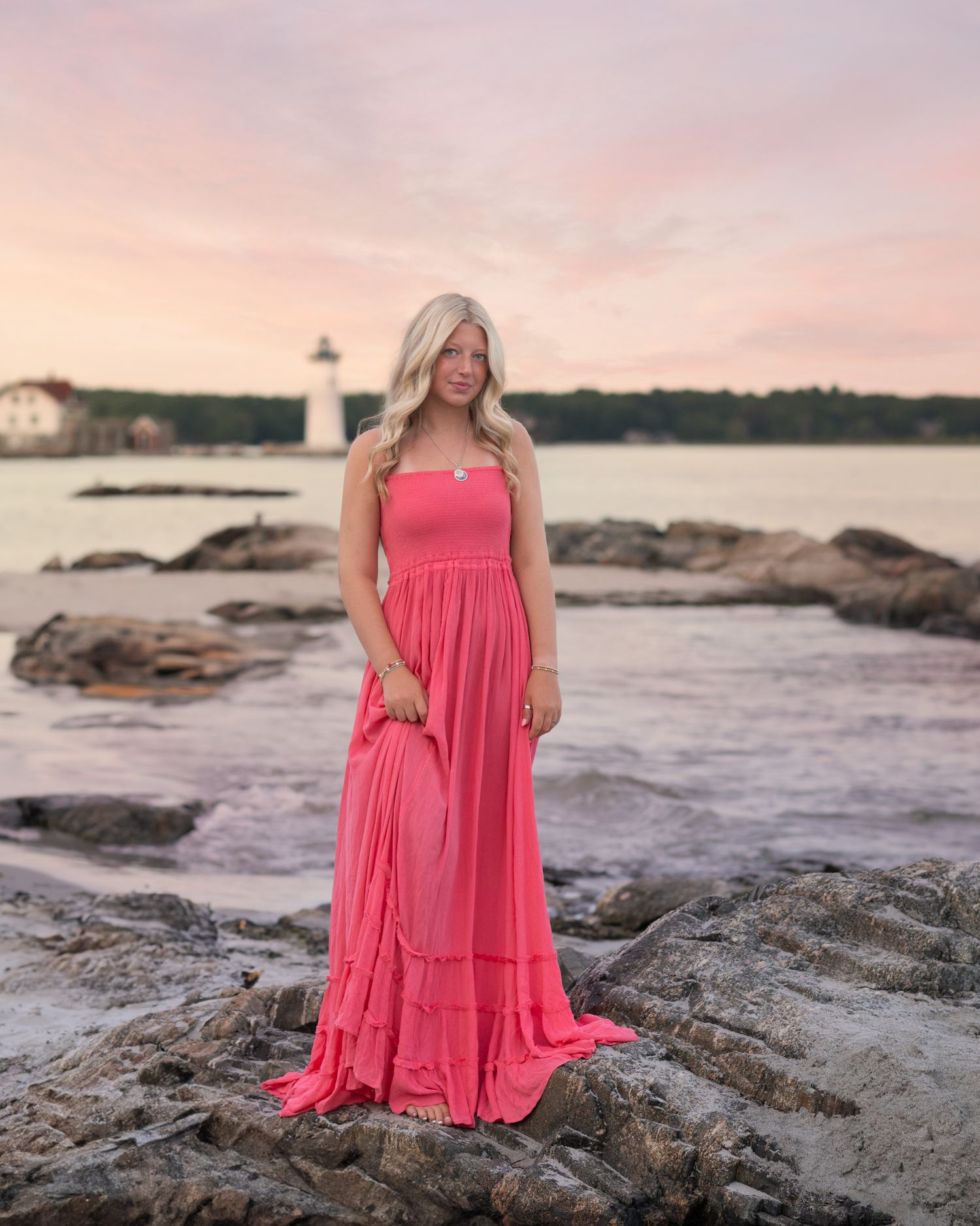 Person in flowing pink dress poses on rocky beach at sunset.