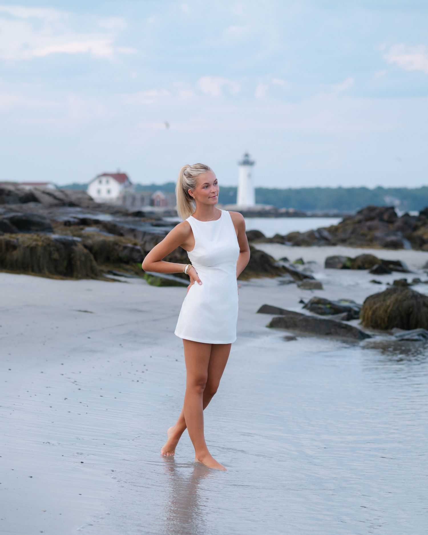 Blonde woman in white mini dress posing barefoot on rocky beach with lighthouse in background.