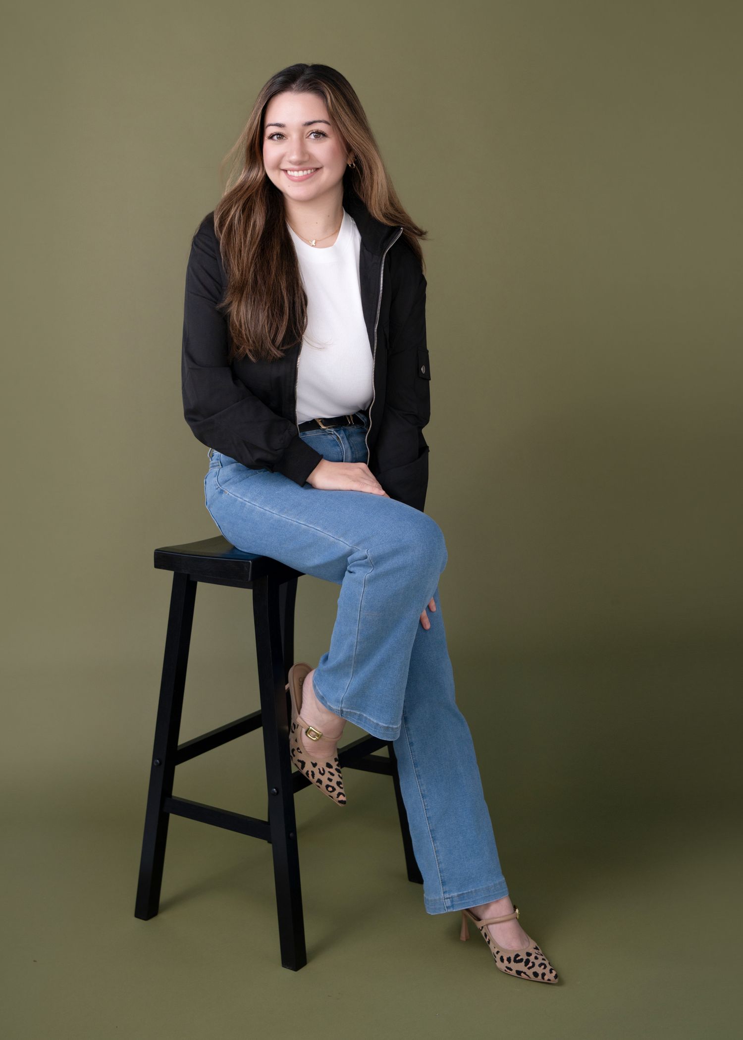 Casual portrait series on black stool wearing jeans and blazer against olive background.