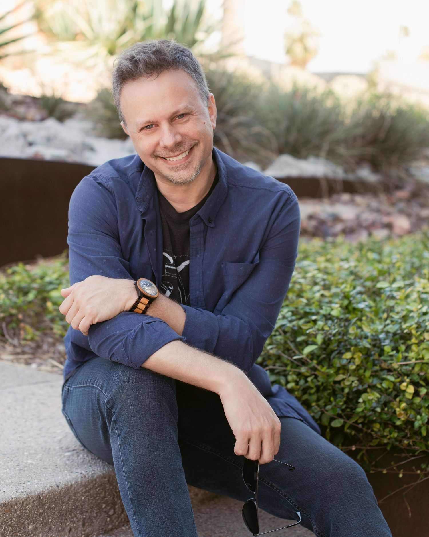 Man in navy blazer sitting casually outdoors against landscaped background.