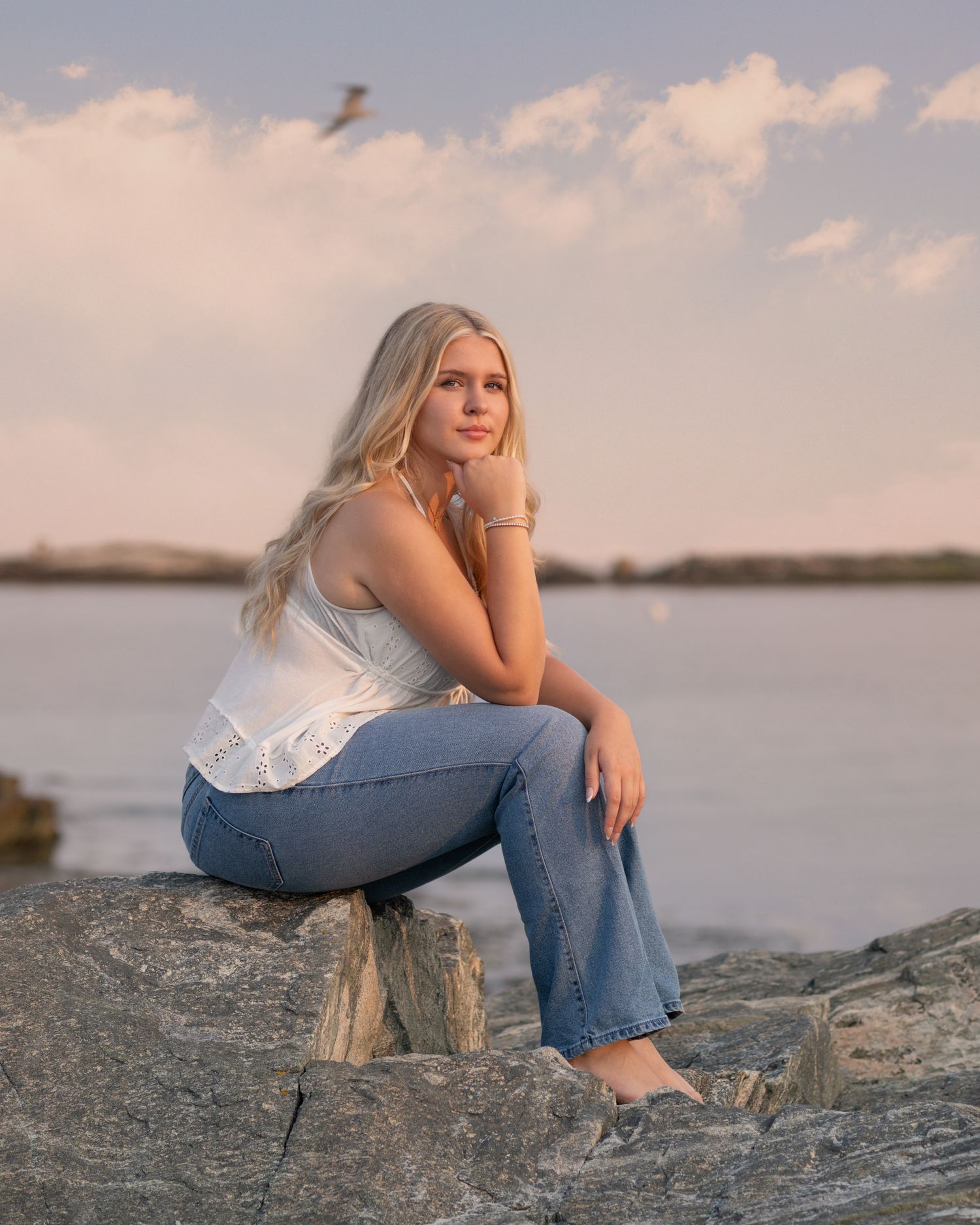 Casual coastal portrait on rocky shore at sunset with water and clouds in background.