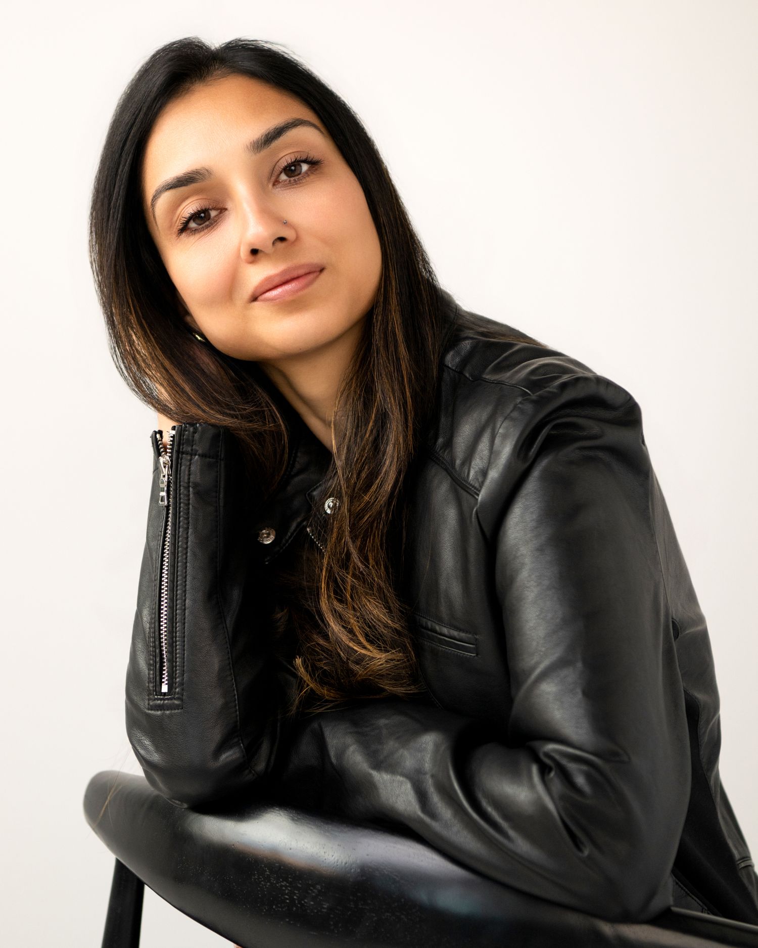 Woman in black leather jacket sitting on chair, long dark hair, posing for portrait photo against white background. Amesbury, MA