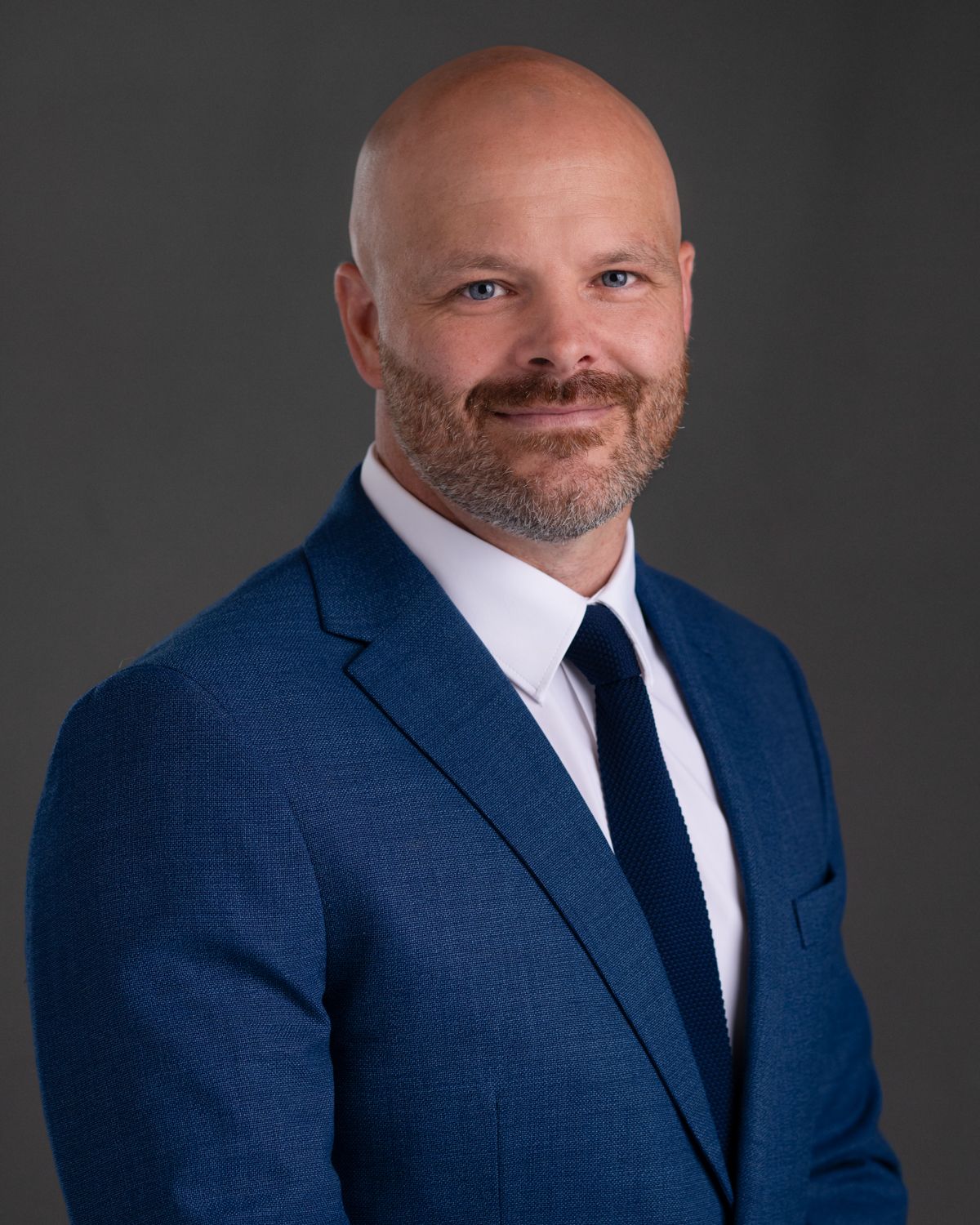 Professional headshots of a person in a navy suit and tie against a dark backdrop.