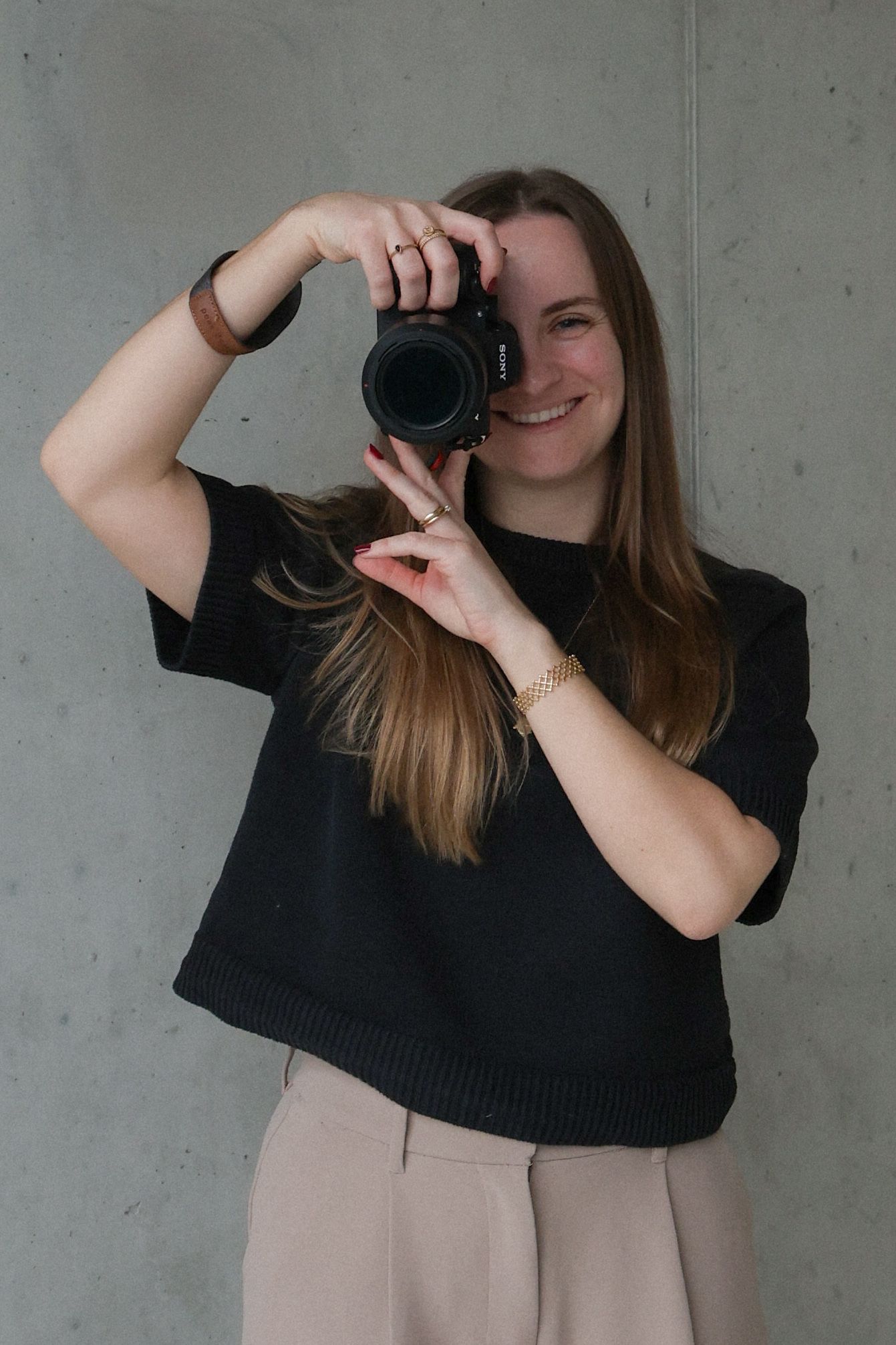 Eine Fotografin in schwarzem Pullover und beiger Hose zeigt verschiedene Kamerahaltungen vor einer grauen Wand.