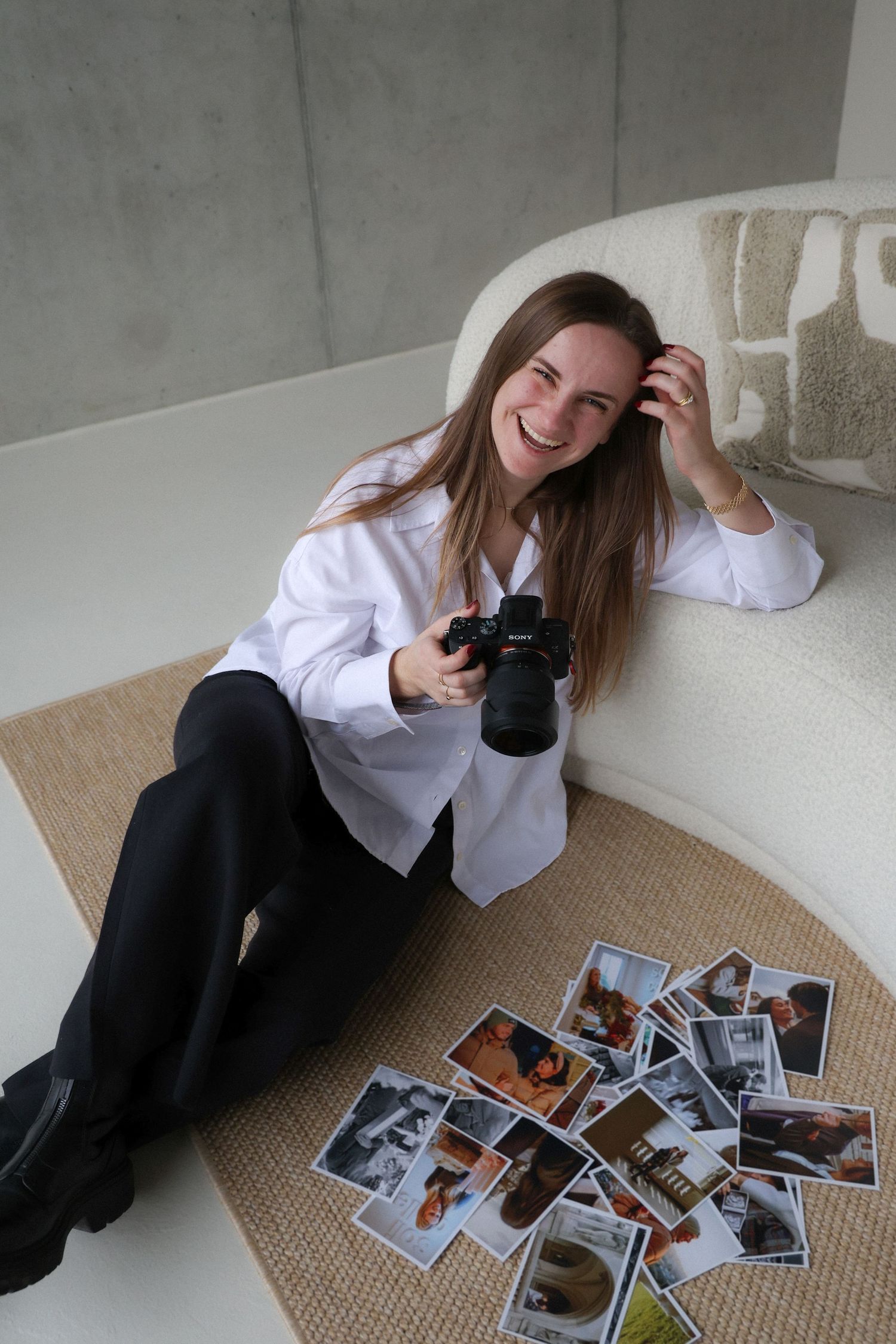 Fotografin im weissen Hemd sitzt auf dem Boden mit Kamera und verstreuten Fotos und lacht beim Durchsehen der Aufnahmen.